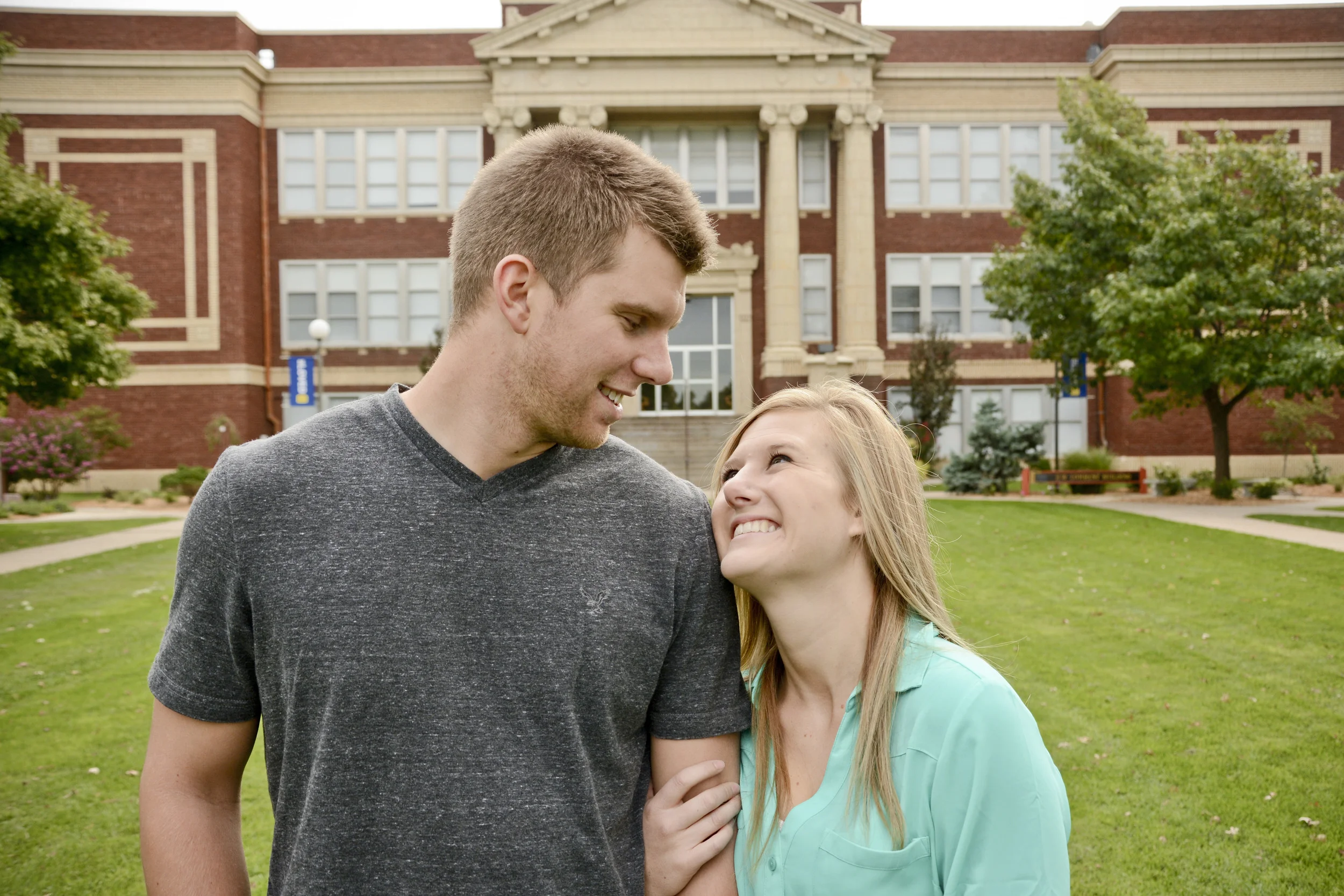 A man and a girl smiling and looking at each other in front of a red brick school building with trees and green grass.