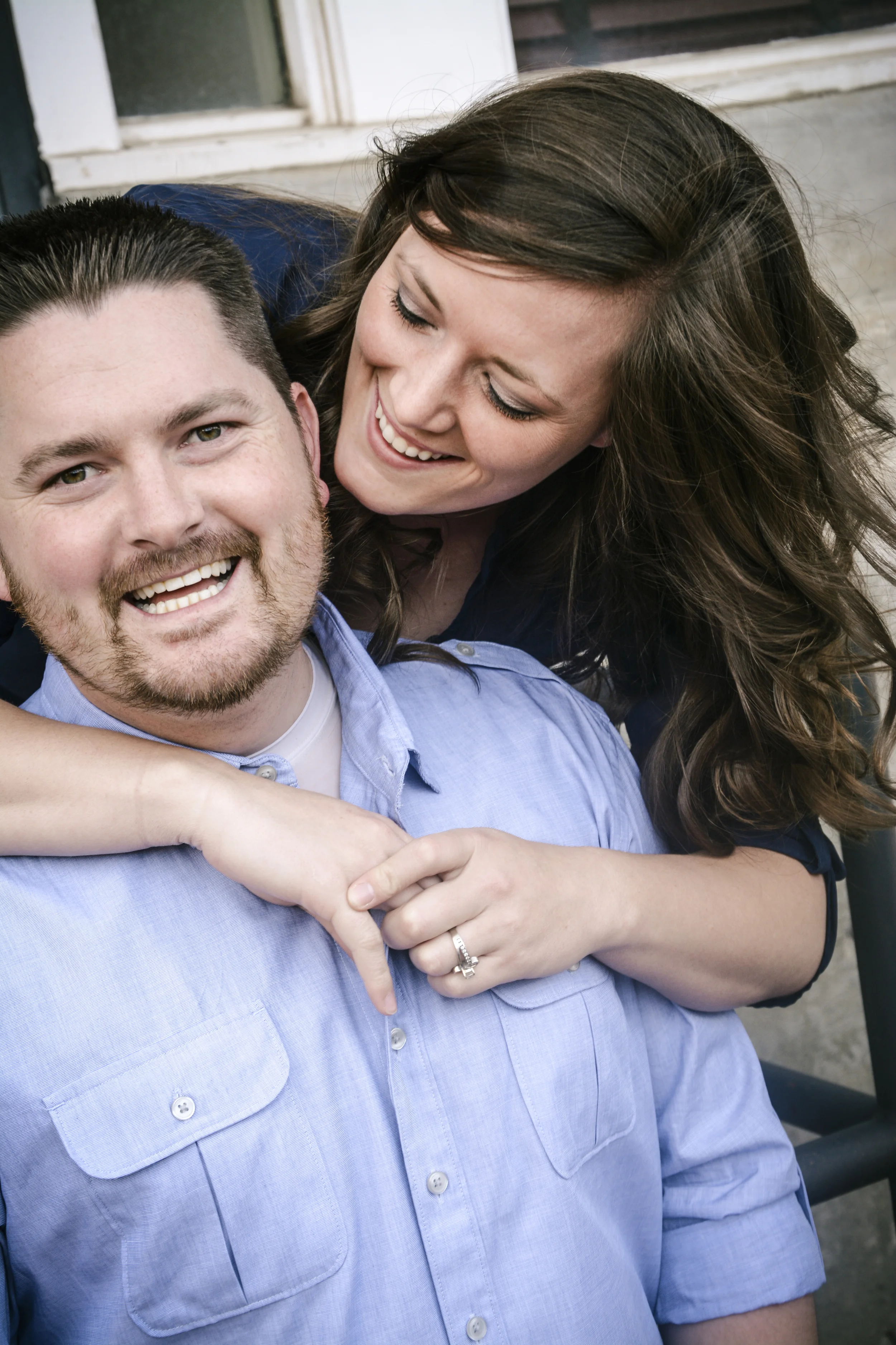 A smiling man with dark hair and a beard wearing a light blue button-up shirt, with a woman leaning over him, smiling and touching his shoulder, with long wavy brown hair and a dark top, outside near a building.