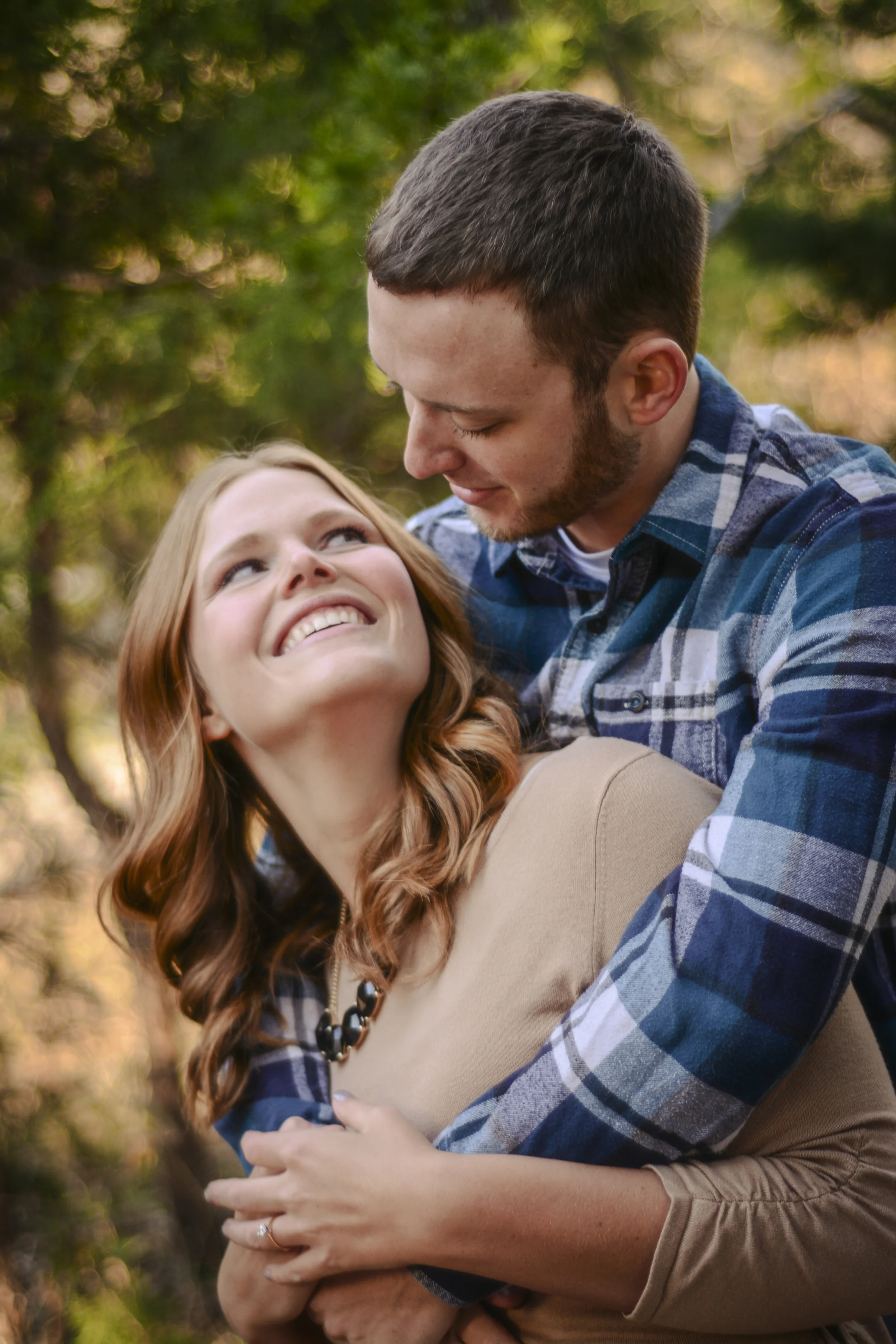 A young woman with red hair smiling up at a young man with short dark hair and a beard, who is looking down at her affectionately while embracing her outdoors in a wooded area during fall.