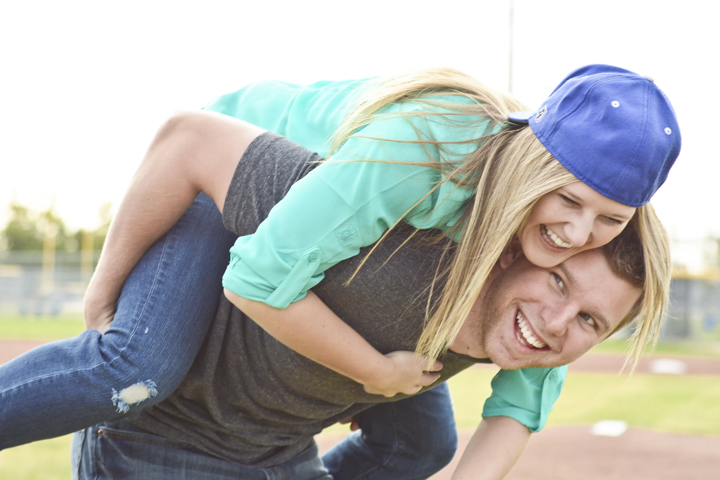 A young woman with long blonde hair wearing a blue cap and teal jacket giving a piggyback ride on a man's back outdoors on a sunny day.