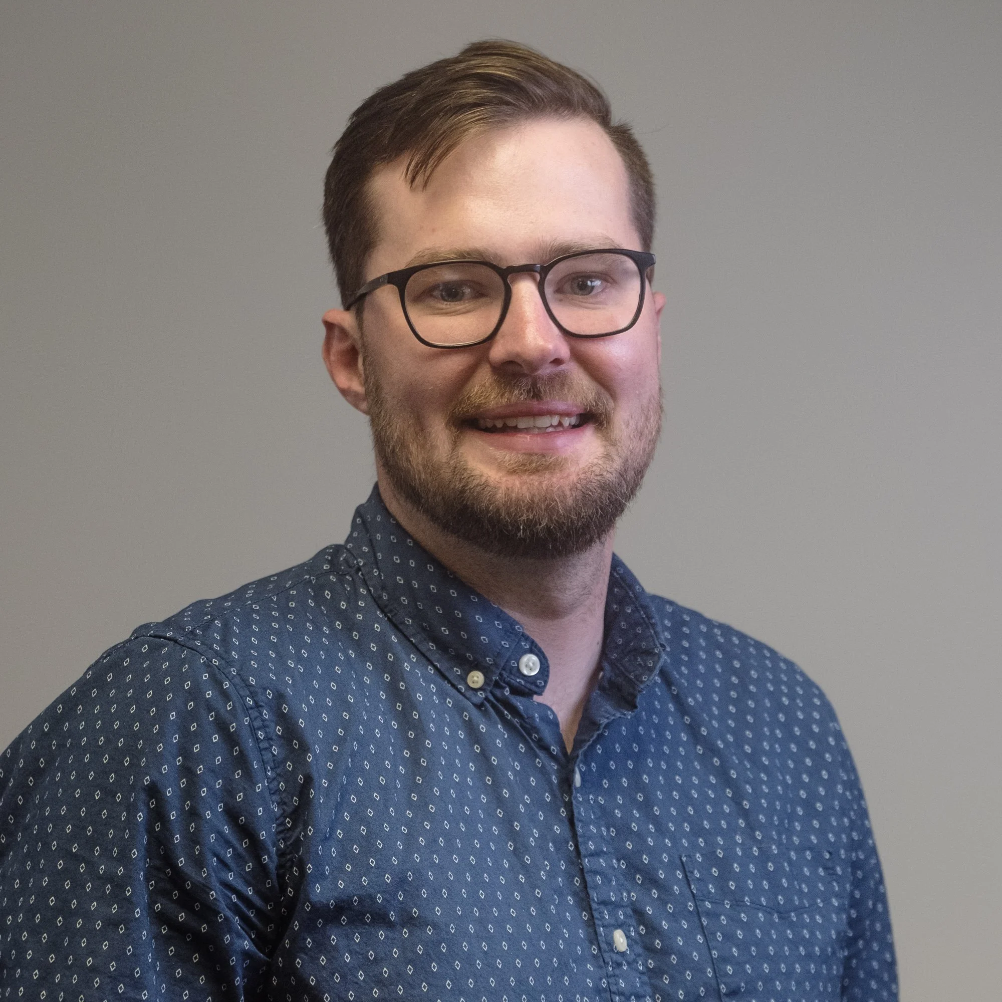 Close-up of a young man with short brown hair, glasses, and a beard, smiling and wearing a dark blue shirt with small white diamond patterns, standing against a plain light gray background.