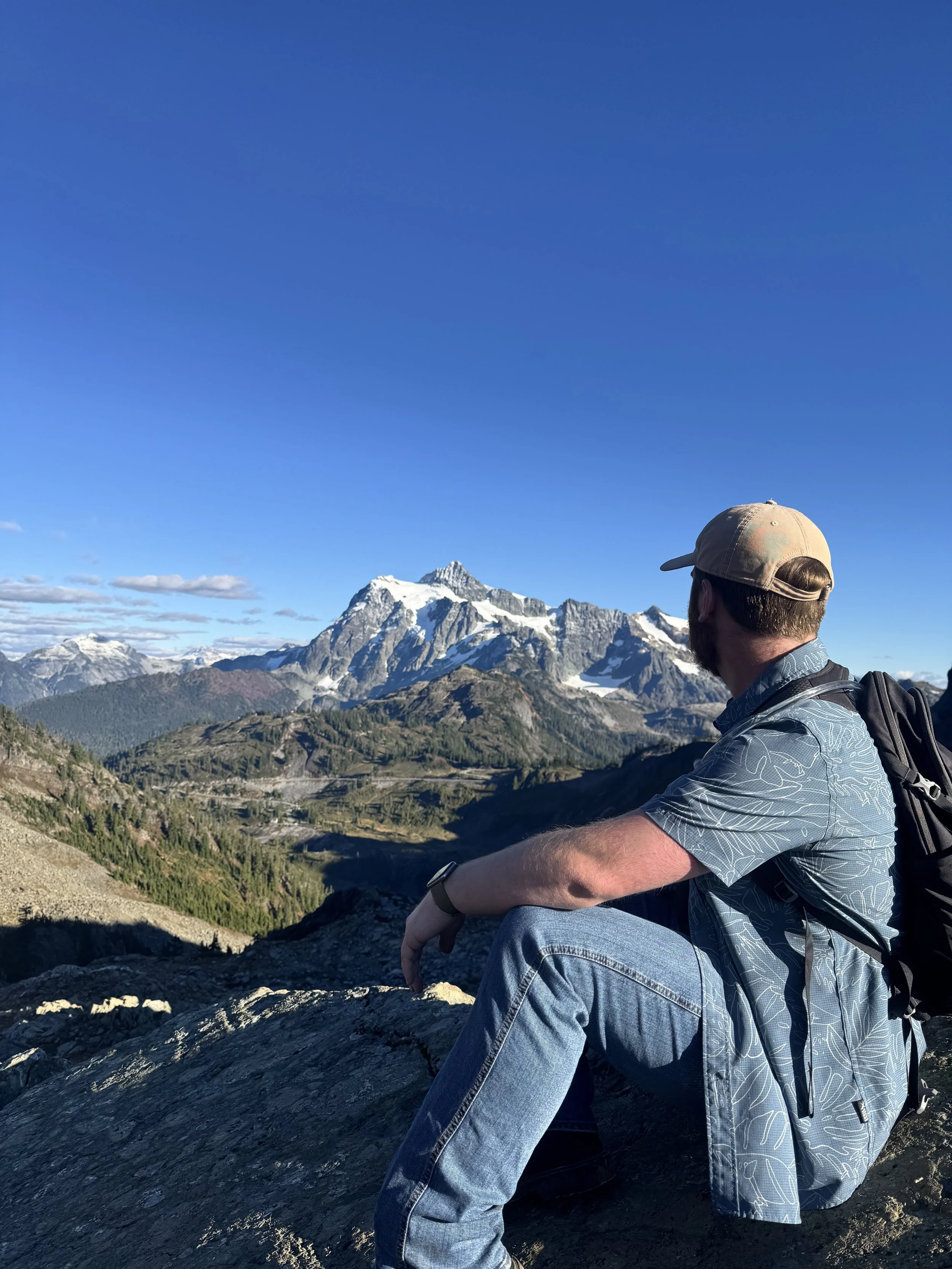 Hiker wearing a cap and backpack sits on a rocky overlook above a forested mountain valley with snow-capped peaks.
