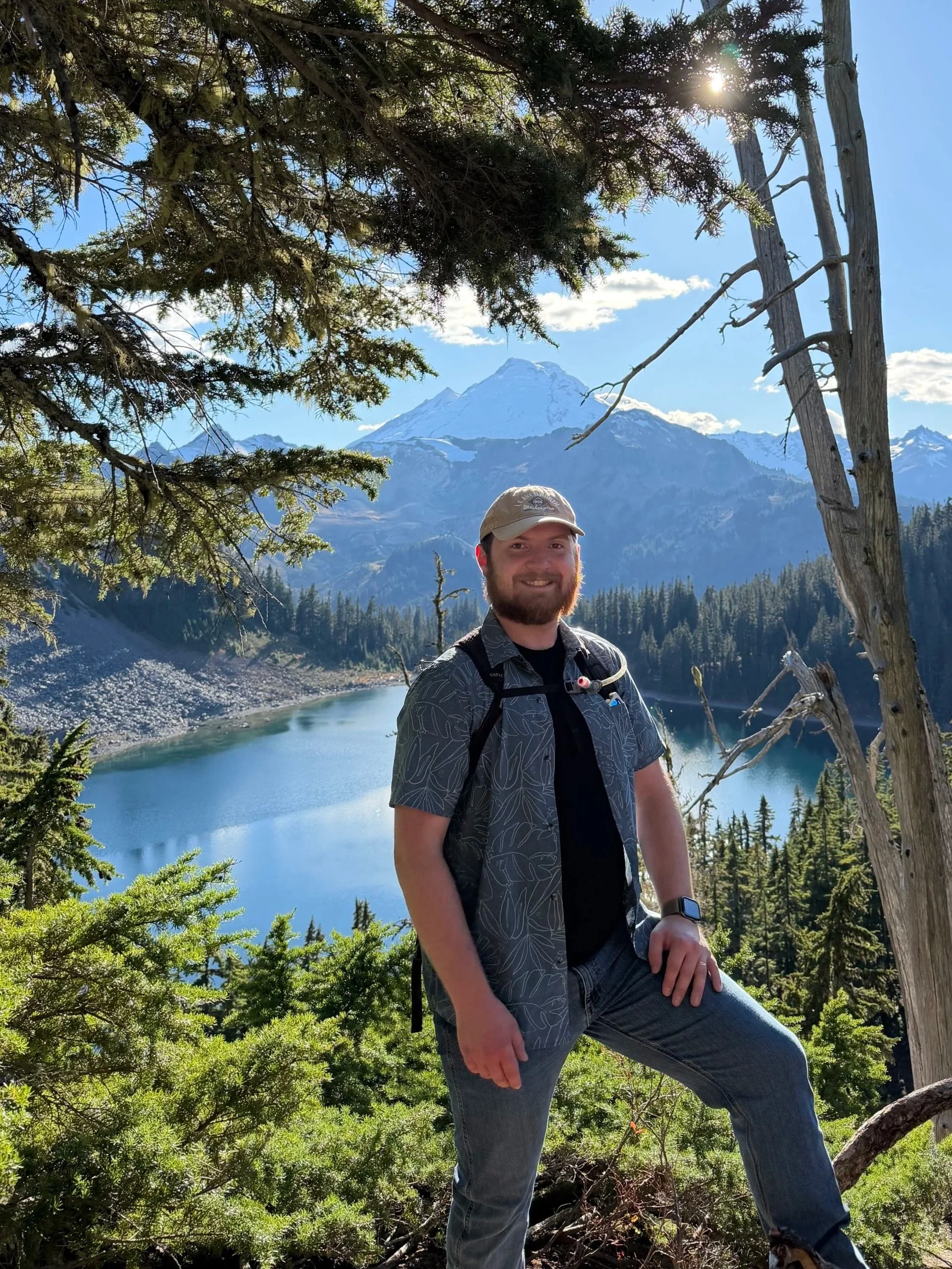 Hiker posing beside a mountain lake with a snow-capped peak and evergreen forest in the background.