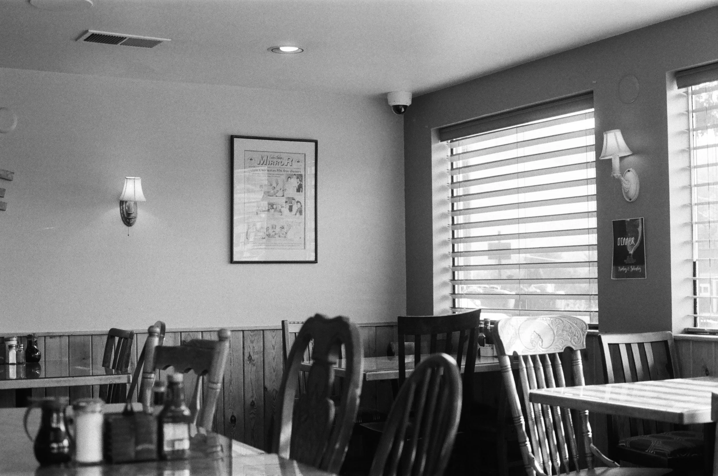 Black-and-white photo of a cozy restaurant dining area with wooden tables, chairs, window blinds, wall lamps, framed artwork, and a security camera.