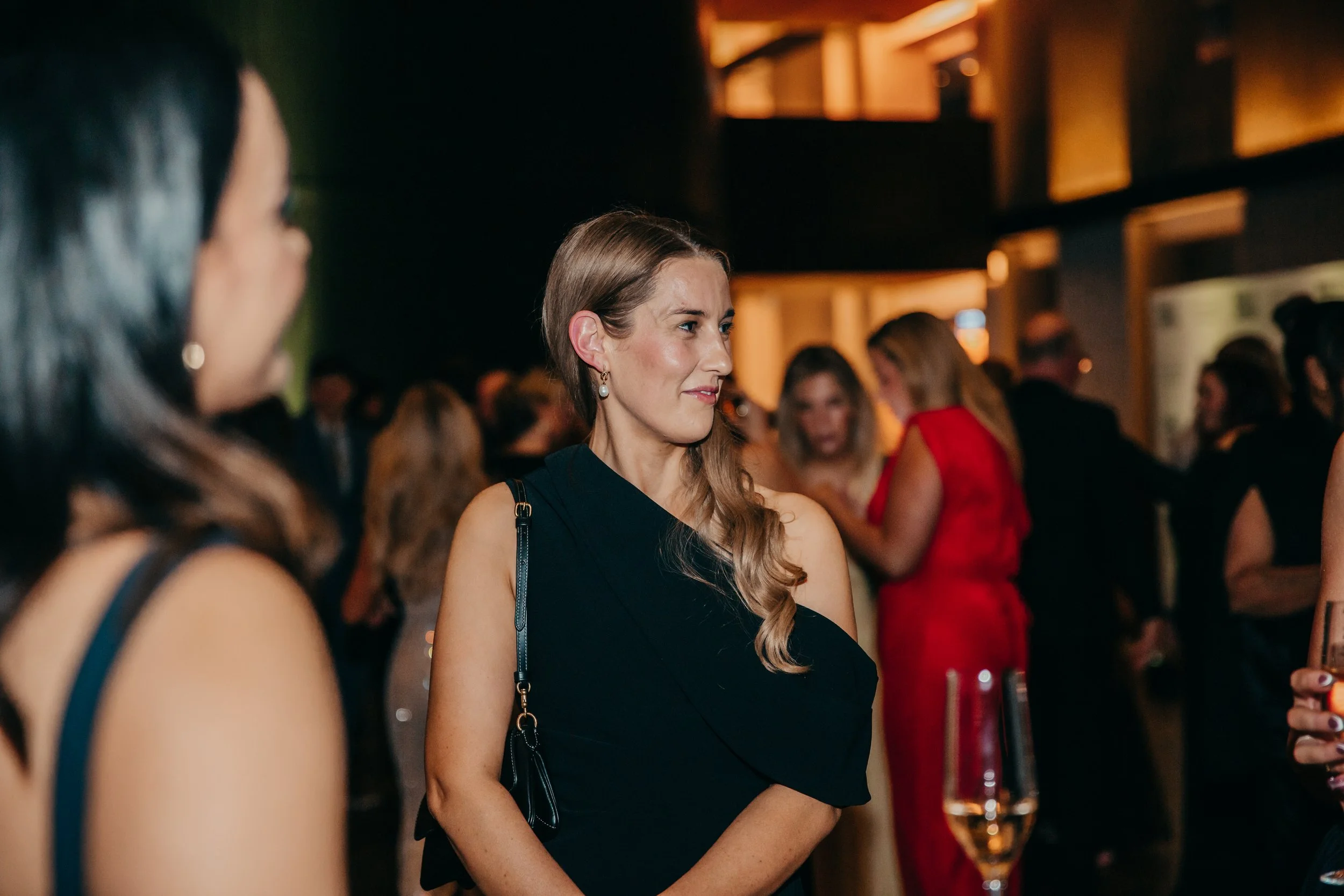 Woman in a black dress at a warmly lit evening cocktail party surrounded by guests