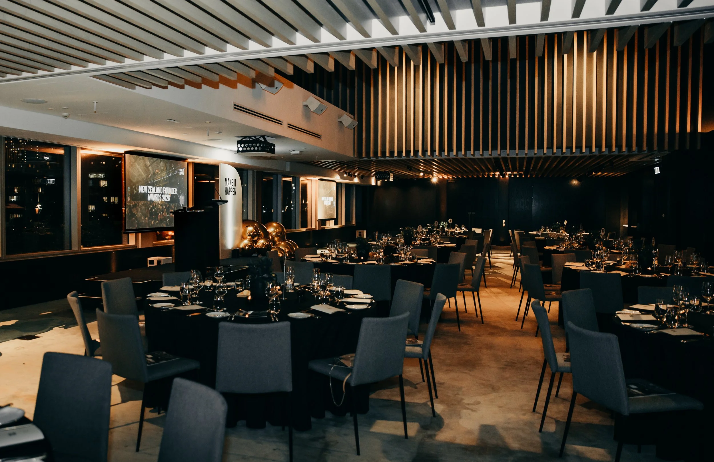 Elegant event venue with round banquet tables, place settings, stage, projector screens, and modern wood-slat ceiling