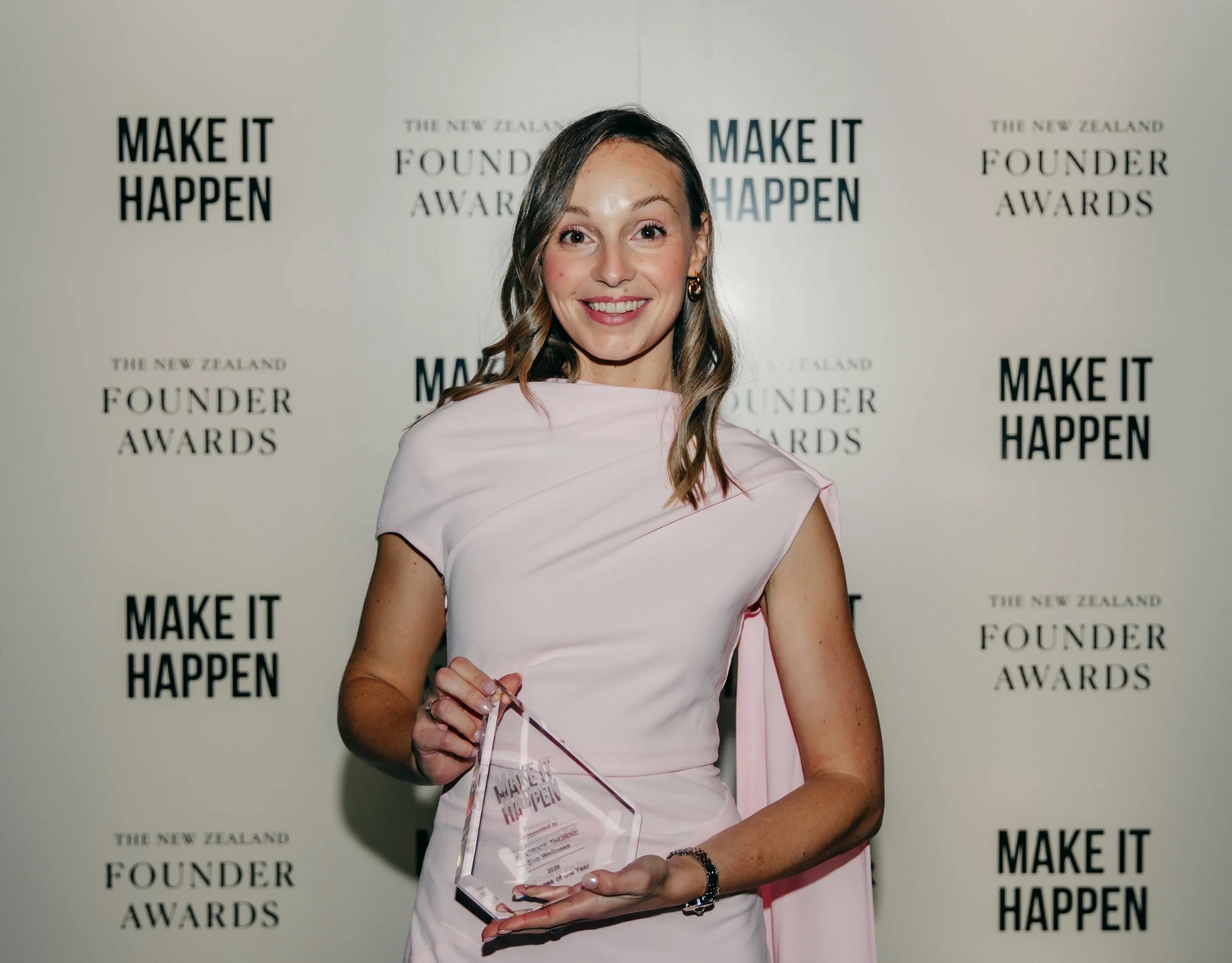 Smiling woman in a pink dress holding a clear Make It Happen award at the New Zealand Founder Awards