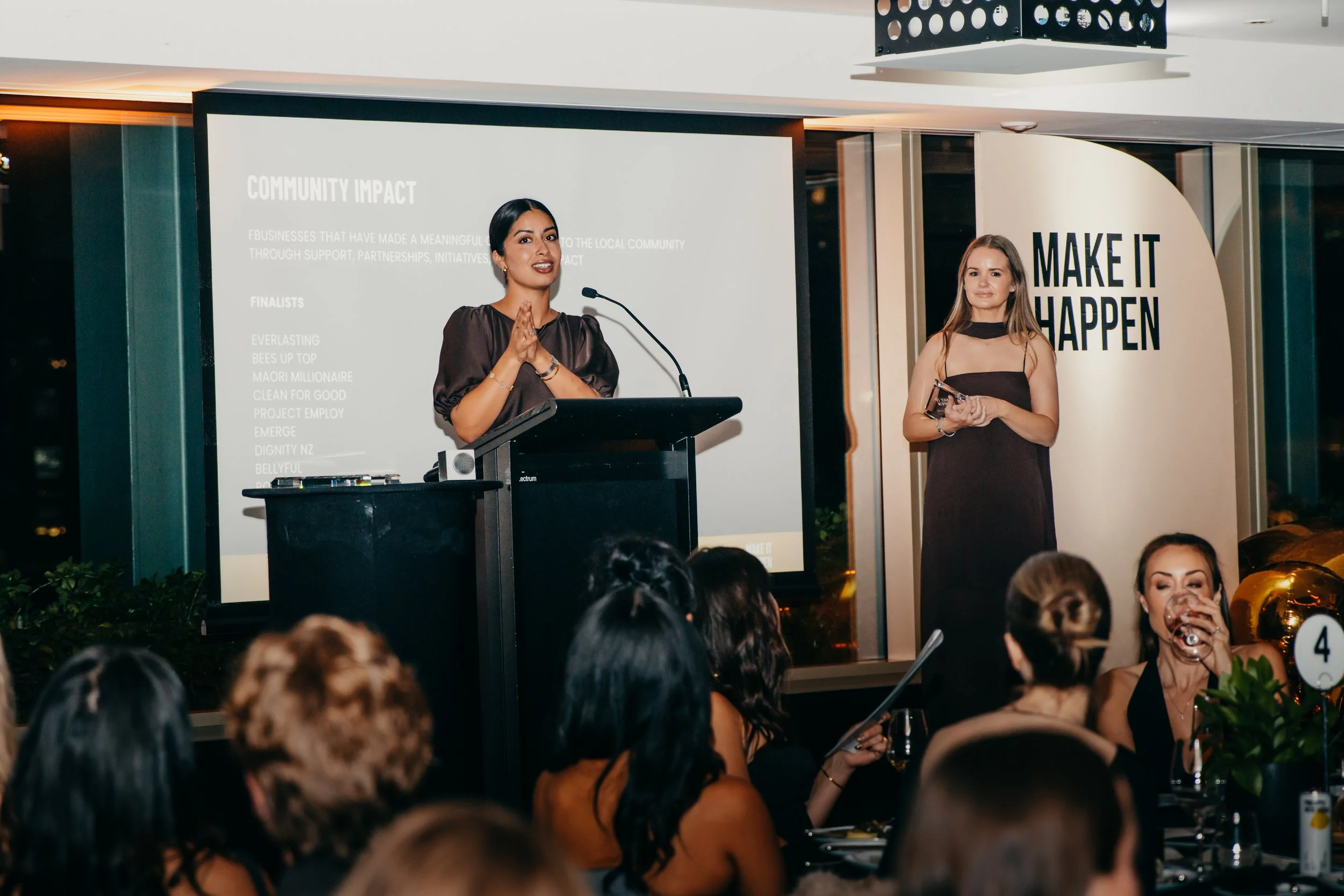 Woman speaking at a podium during a community impact awards event as an audience watches.