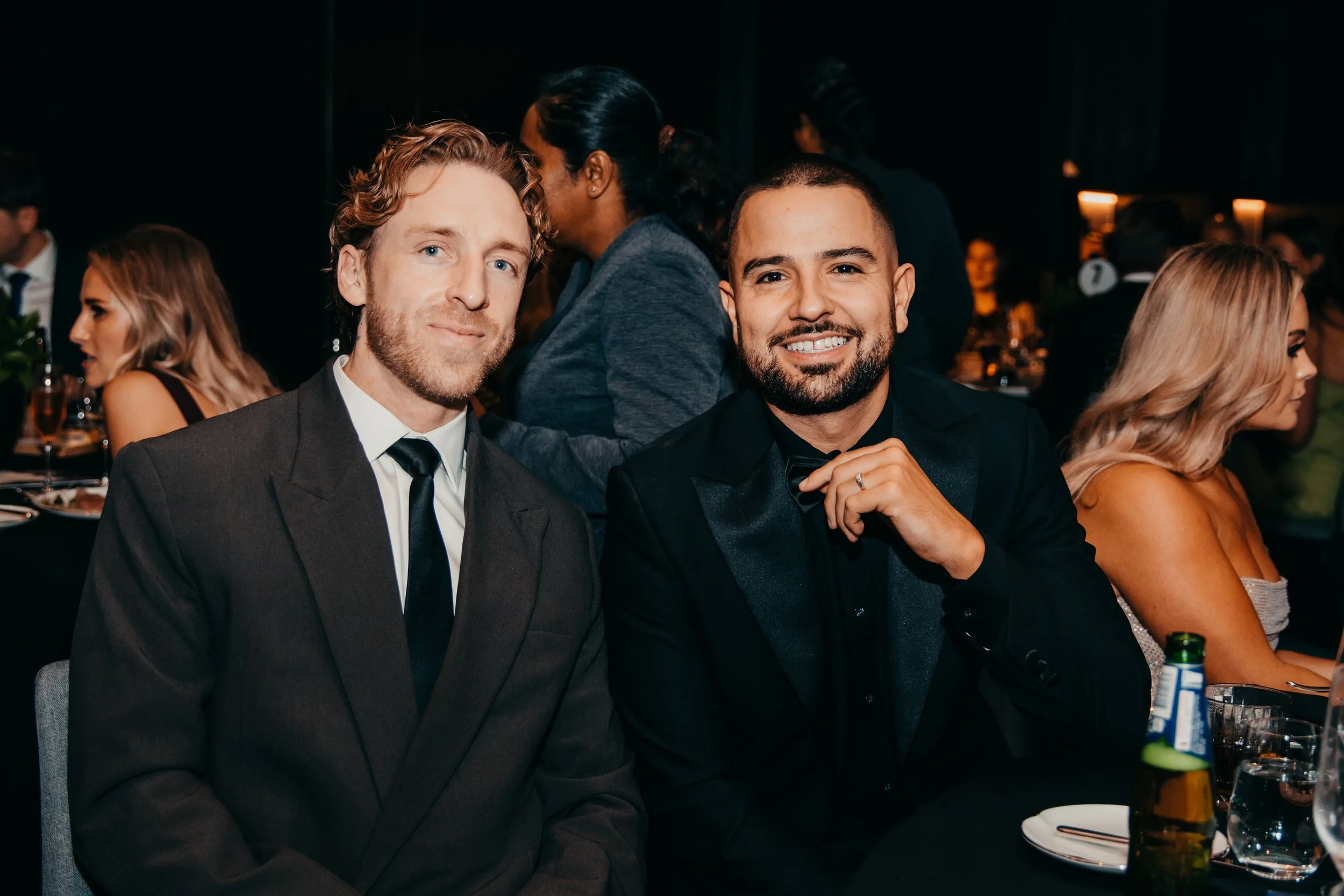 Two men in formal suits smiling at a dimly lit dinner event, with guests and drinks in the background.