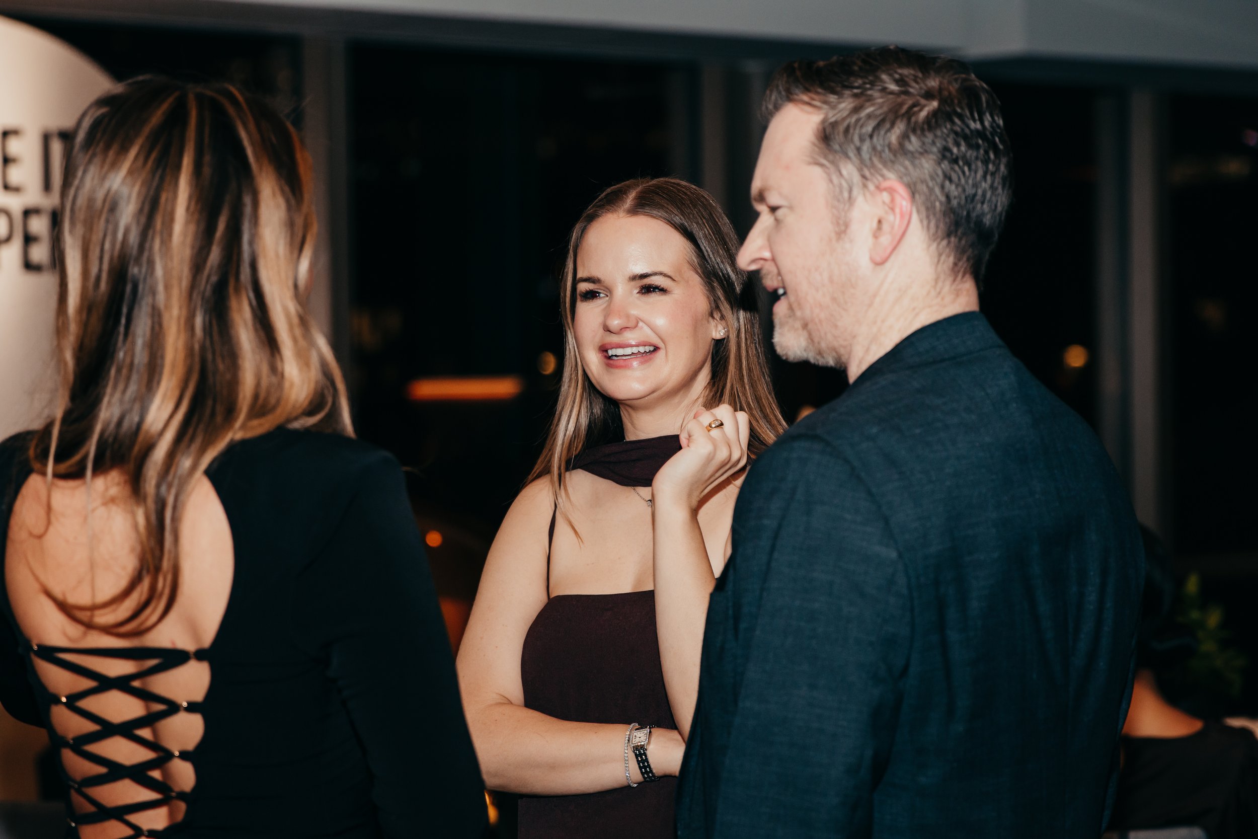 Three people socializing at an evening event, with a smiling woman chatting beside a man in a dark jacket.