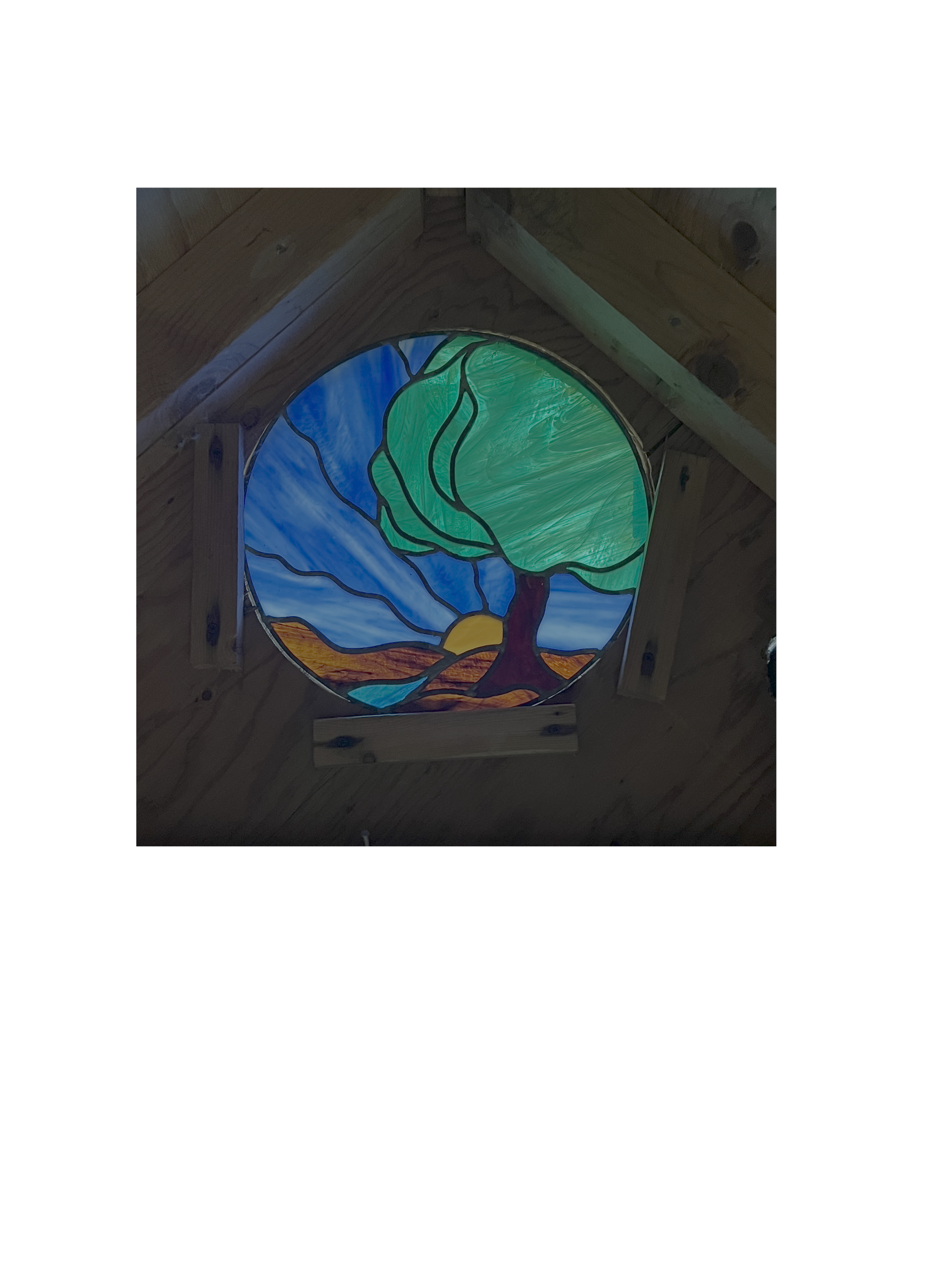 Colorful stained-glass window depicting a large green tree beside a blue river and sunset landscape in a wooden wall
