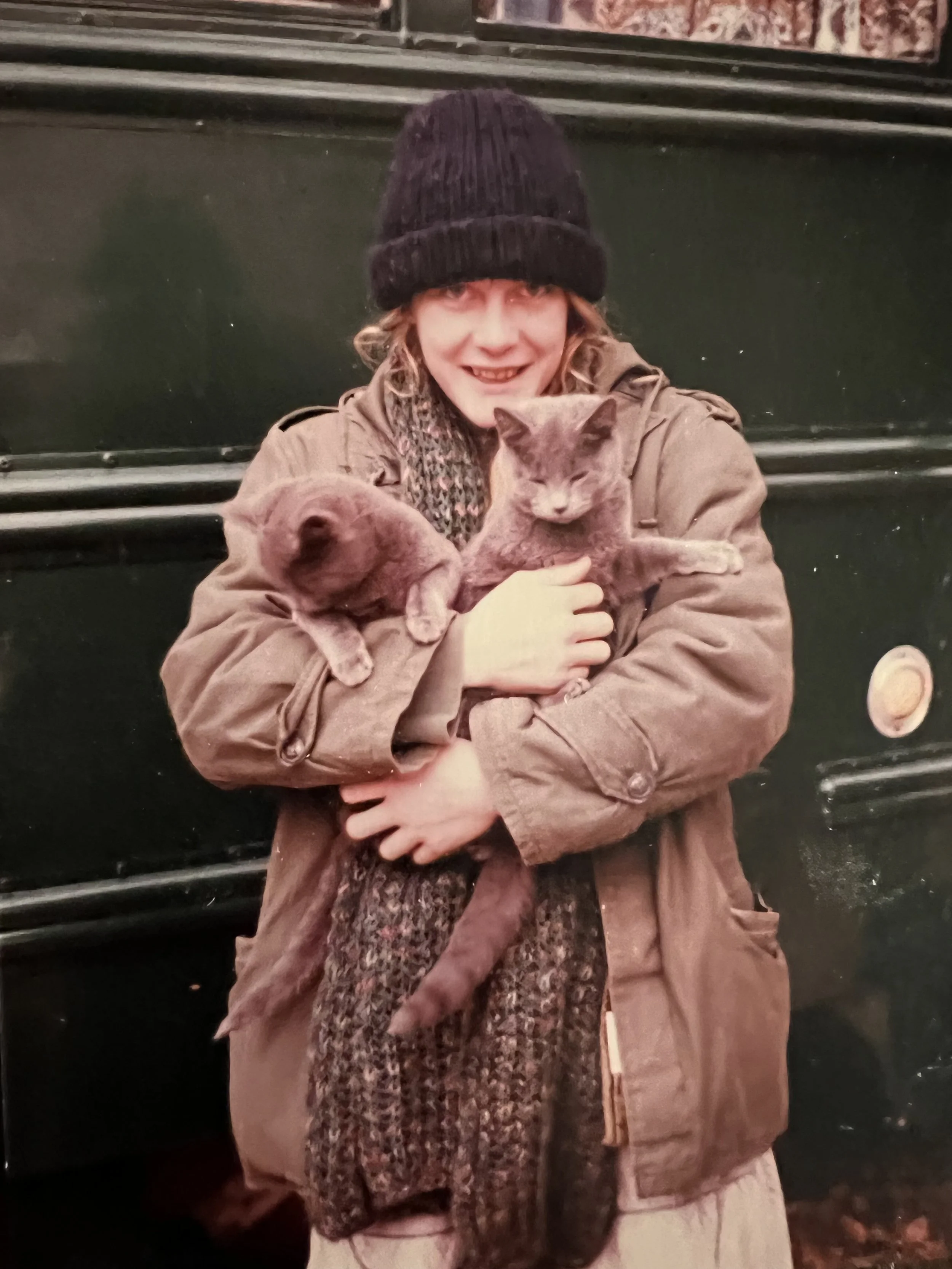 Smiling person in a black knit hat and scarf holding two cats in front of a dark bench.