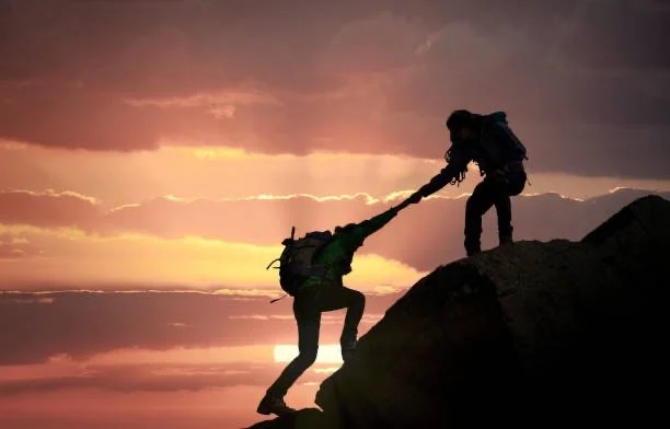 Silhouetted hiker helping another climber up a rocky mountain ridge at sunset