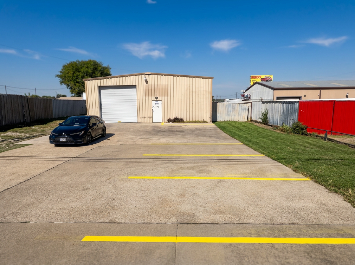 A small parking lot with yellow lines, one black car parked on the left, and a beige warehouse building with a white garage door and a small tree in front. There is a grassy area with bushes and a red fence on the right, under a blue sky with some clouds.