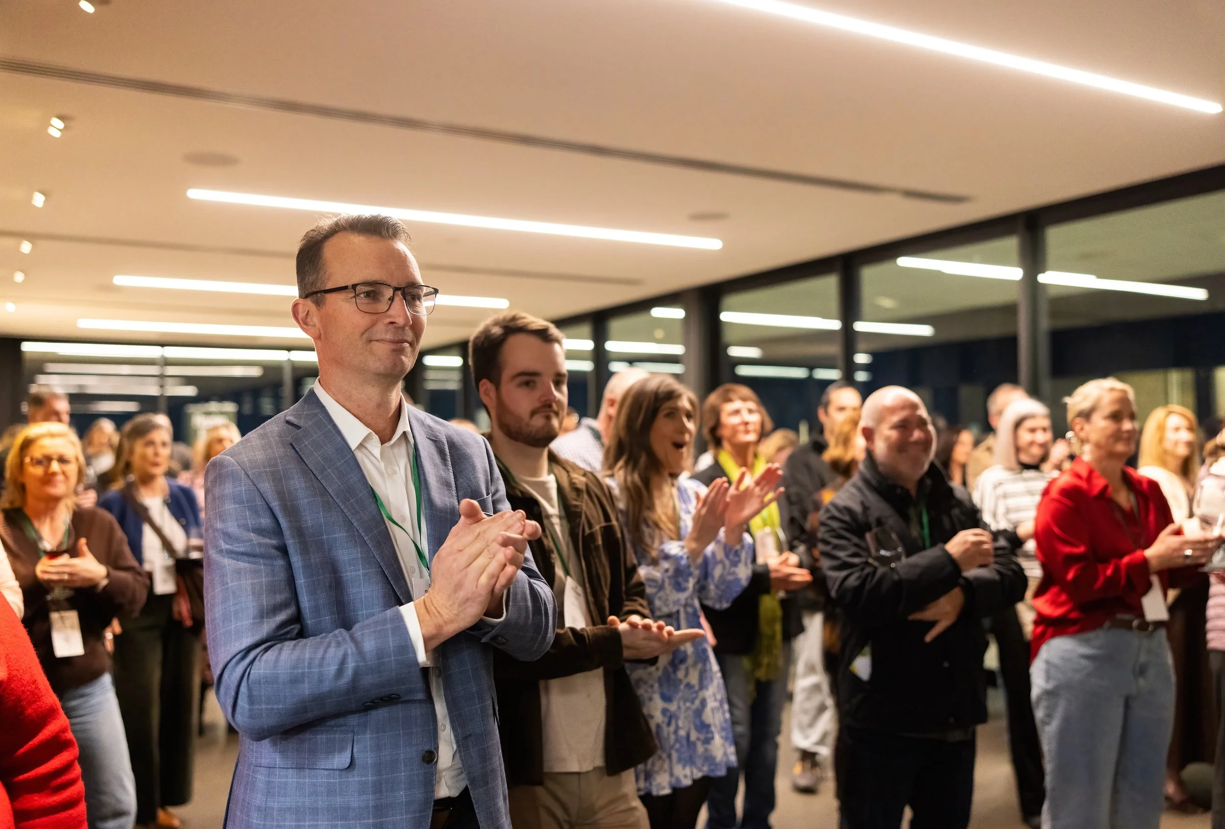 Diverse audience applauding at an indoor business event, with a man in a blue suit in the foreground.