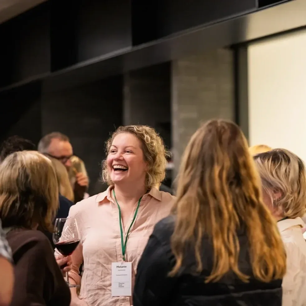 Smiling woman wearing a conference badge and holding a glass of red wine while talking with attendees at an event