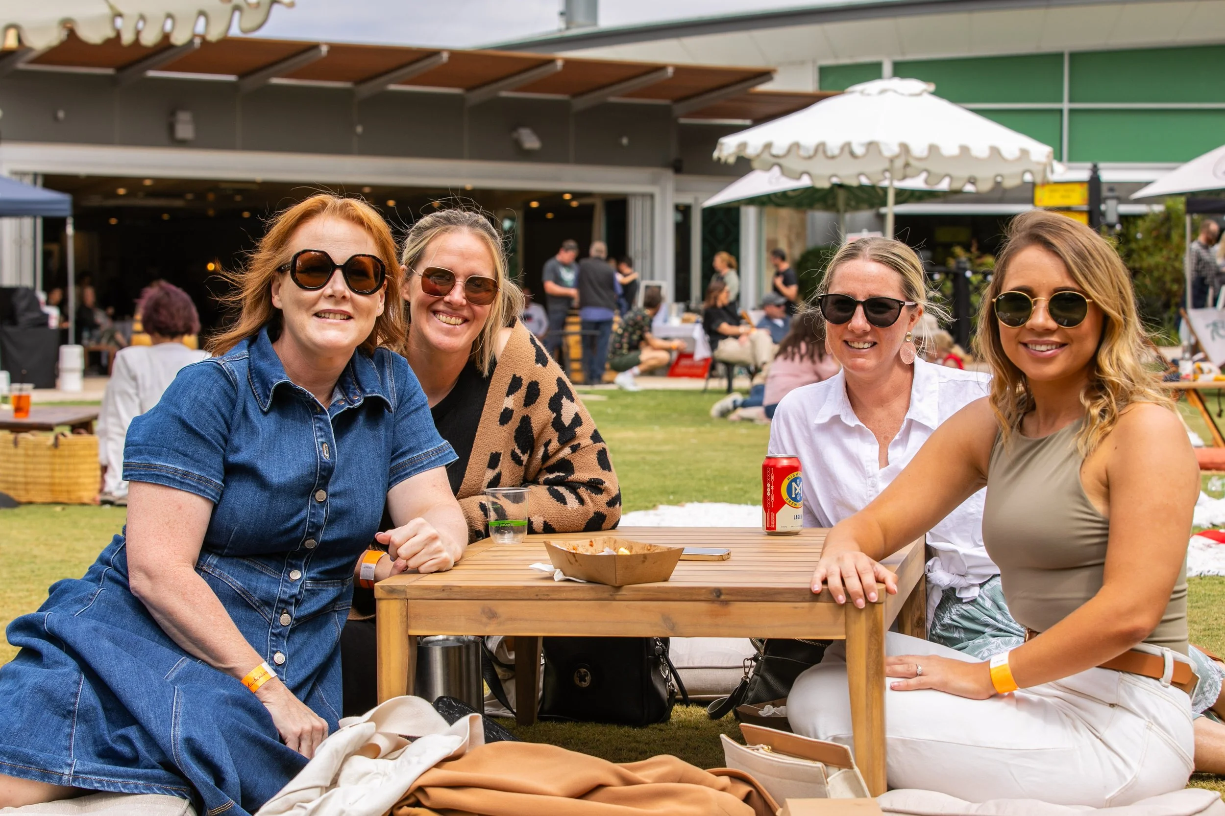 Four women wearing sunglasses socialize at an outdoor café table with umbrellas and drinks.