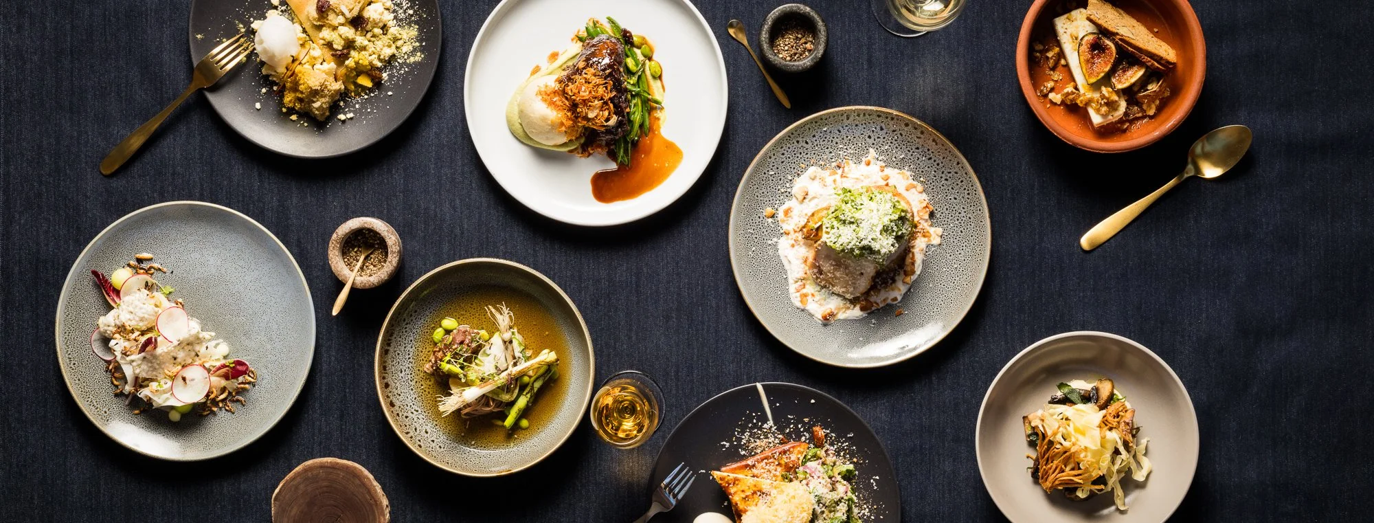 Overhead view of assorted gourmet dishes, desserts, drinks, and tableware arranged on a dark dining table