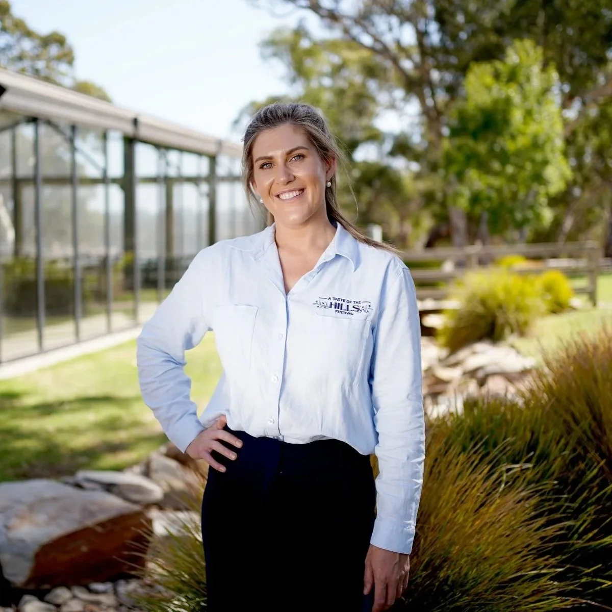 Smiling woman in a light blue festival shirt standing outside a modern glass building in a landscaped garden