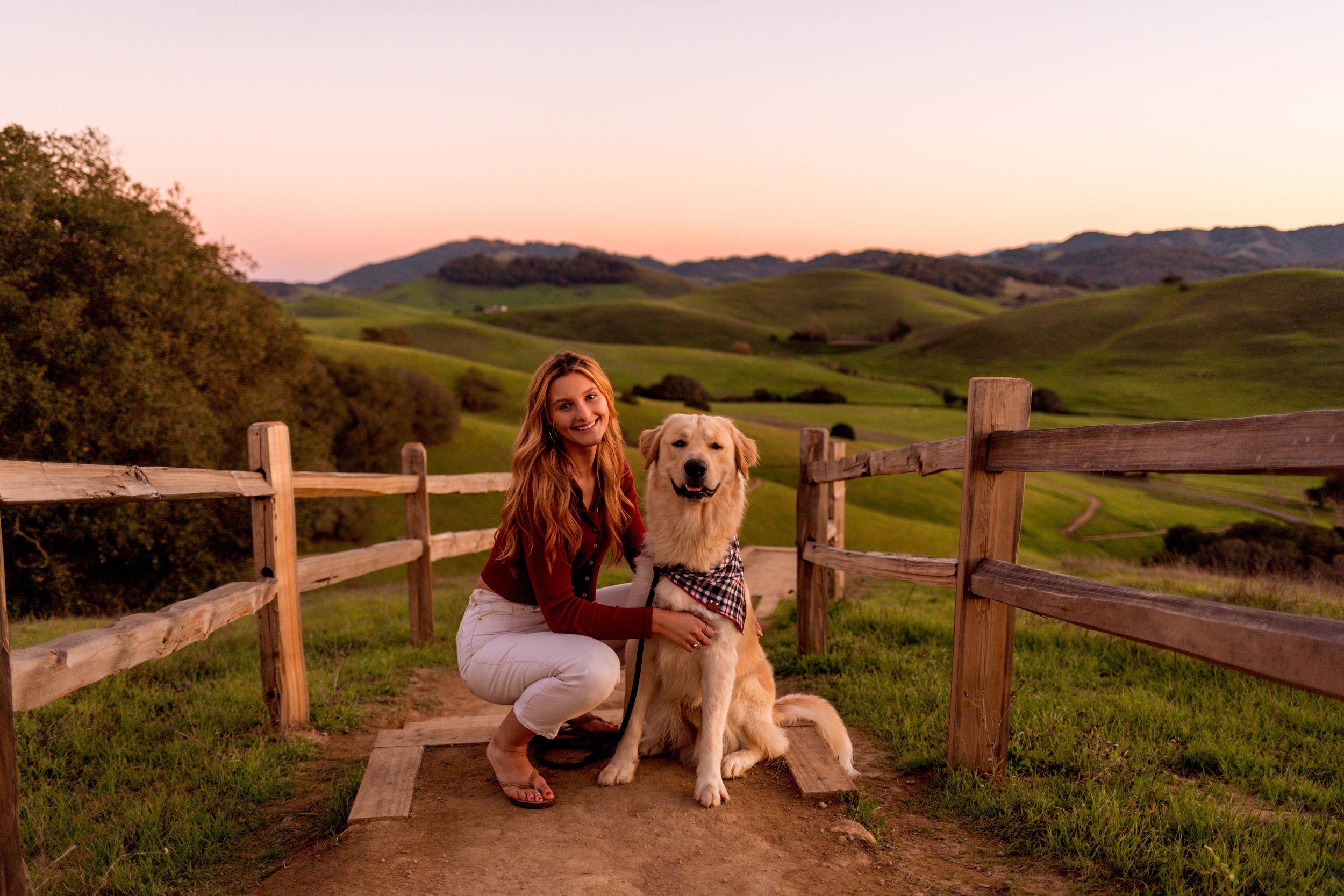 Woman kneeling beside a golden retriever wearing a plaid bandana by a wooden fence in rolling green hills at sunset