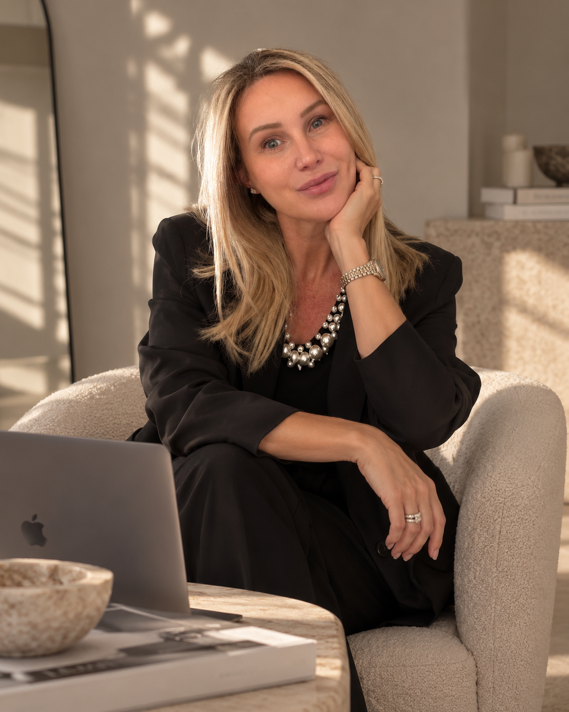 Angela Mckinnon in a black suit and pearl necklace sitting in an armchair beside an open laptop for Socially Now