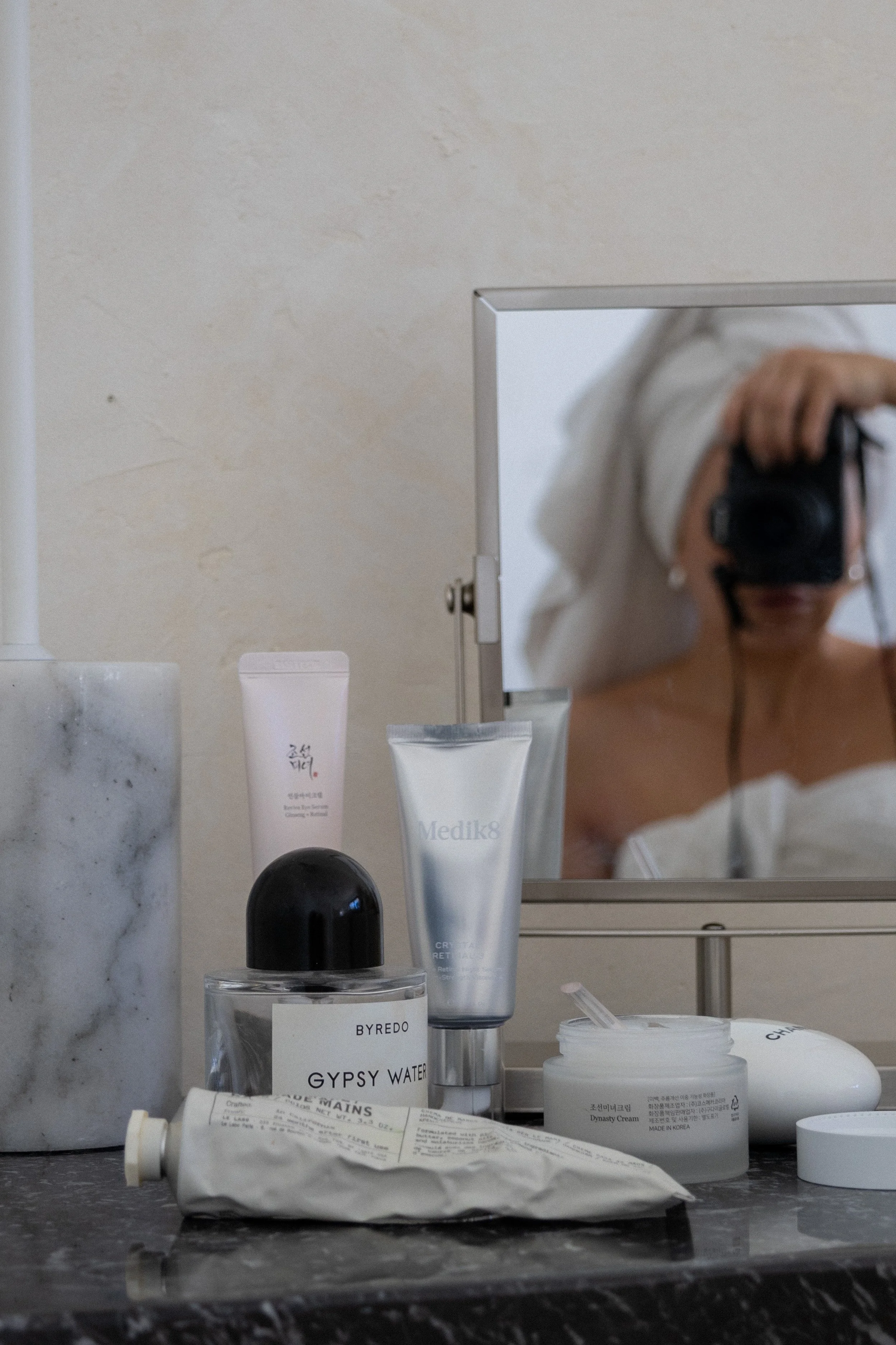Socially Now's skincare products are arranged on a bathroom counter, including Byredo Gypsy Water fragrance, Medik8 cream, and a mirror reflecting a person.