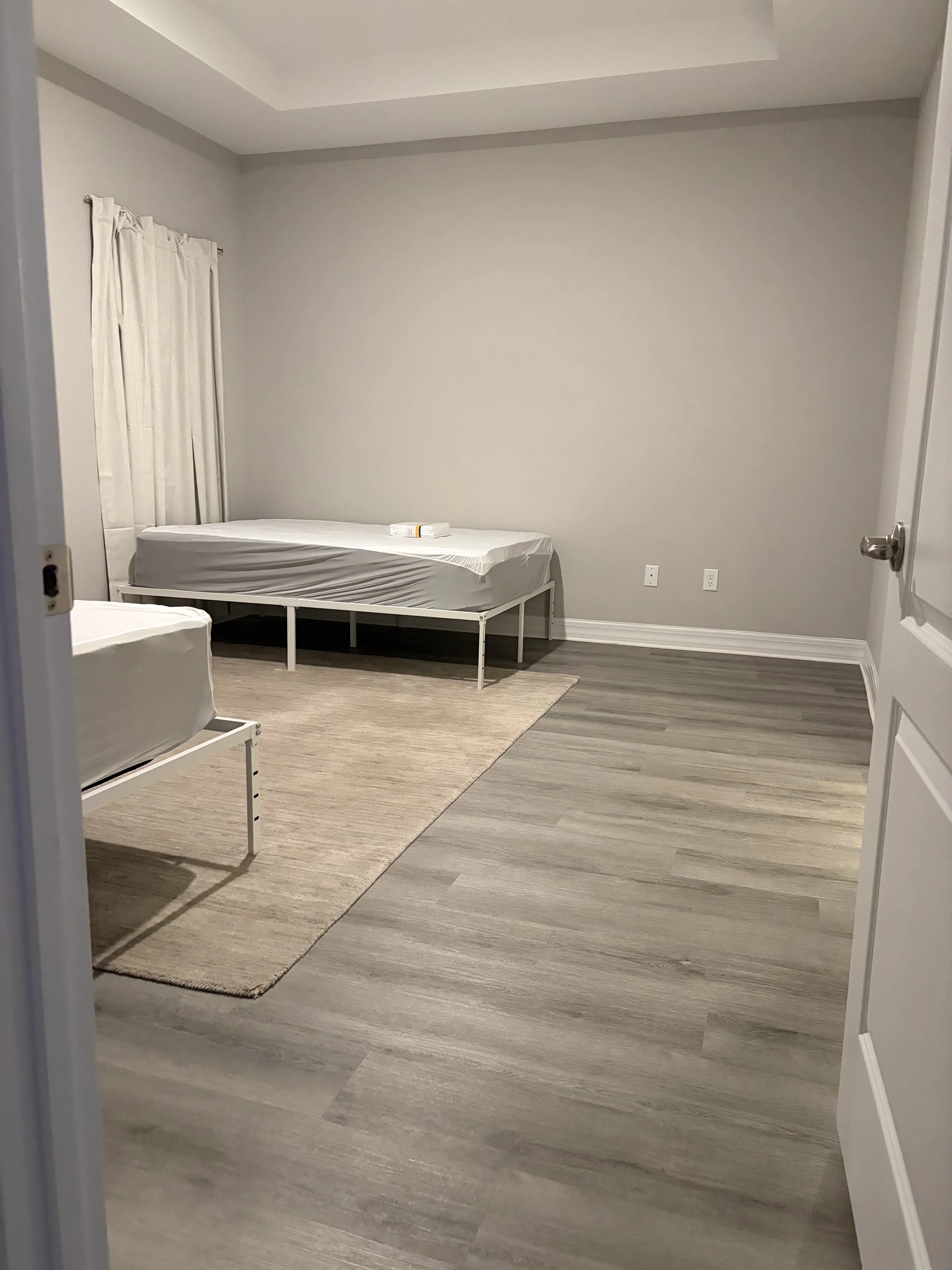 Empty bedroom with a white metal bed frame, mattress, curtains, gray walls, wood-look flooring, and area rug