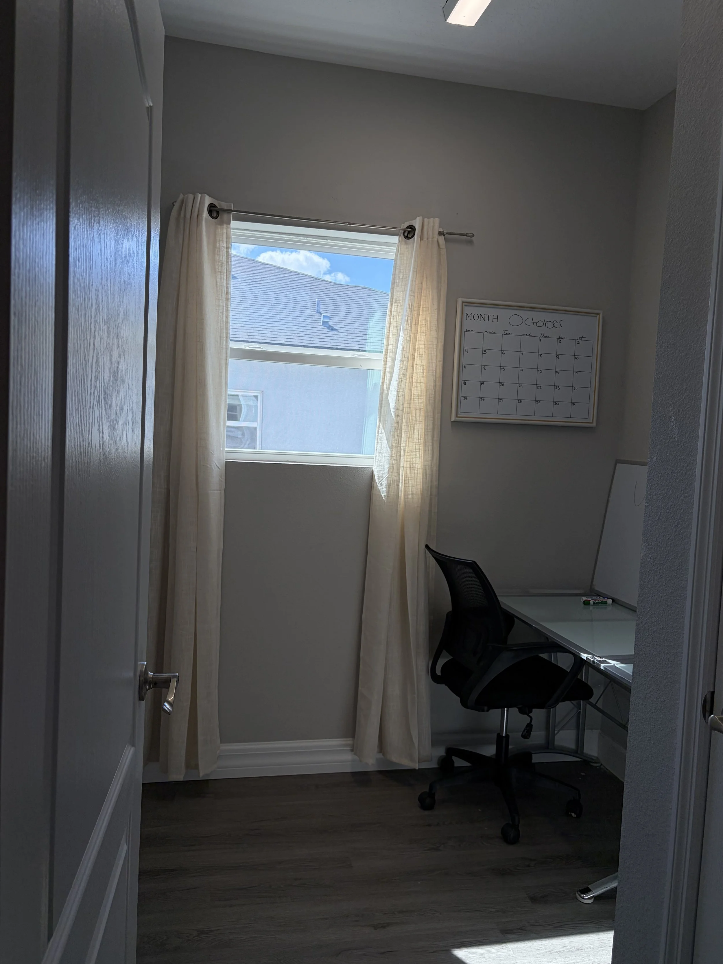 Home office with a curtained window, black office chair, glass desk, and October calendar