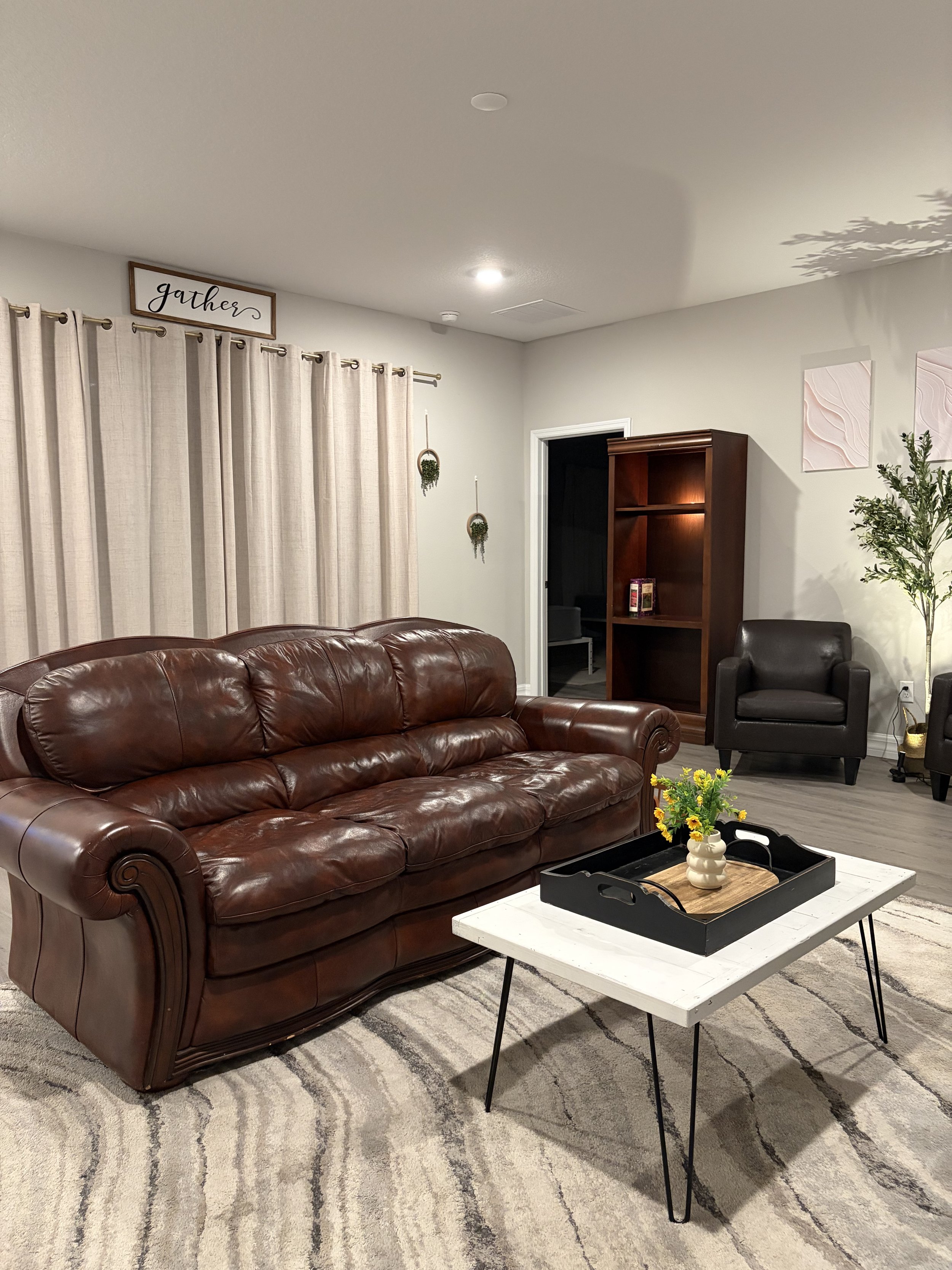Cozy living room with a brown leather sofa, white coffee table, black tray, flowers, armchair, bookcase, curtains, and decorative wall art.