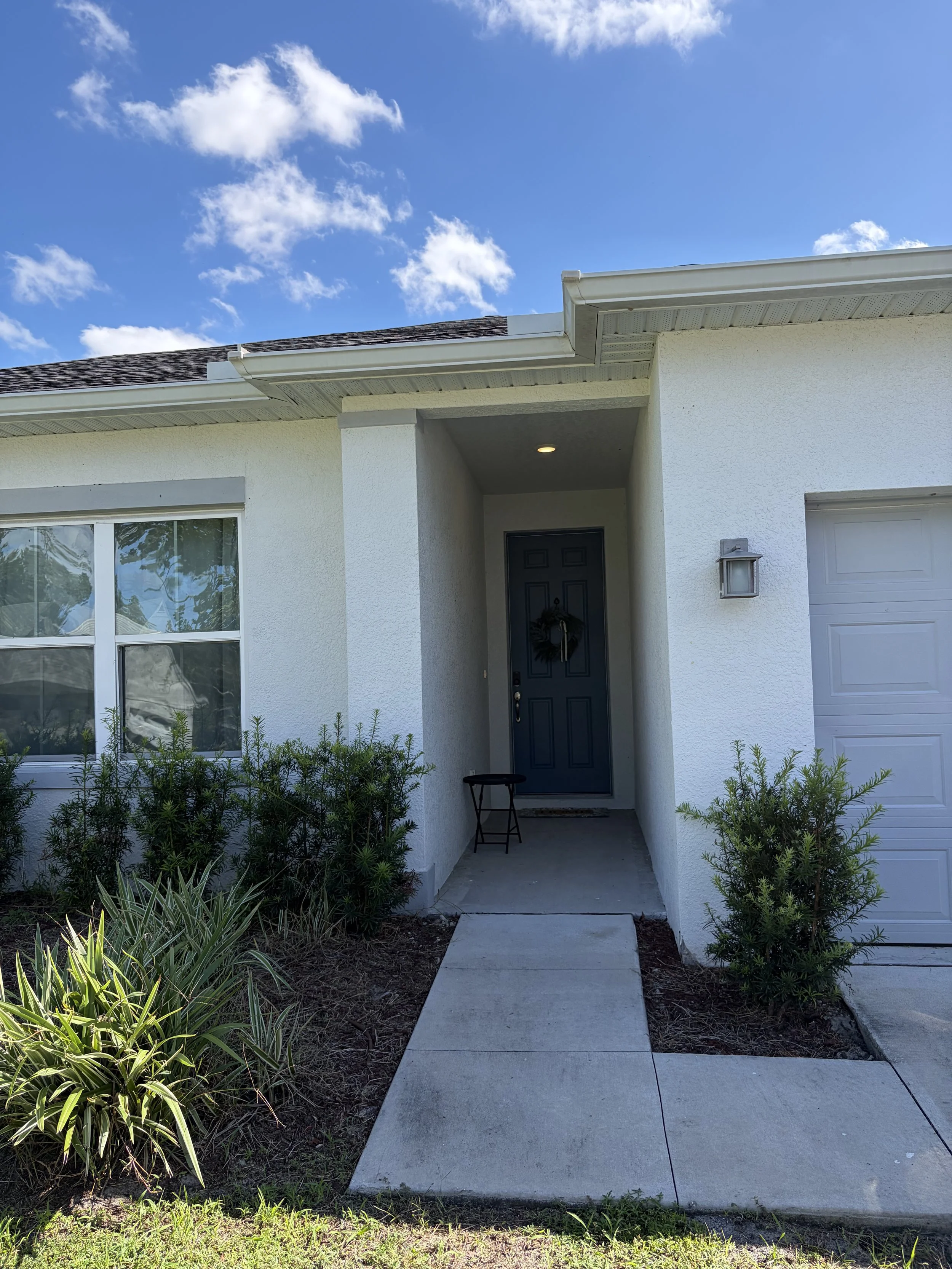 Front entrance of a stucco house with a dark door, garage, walkway, landscaping, and blue sky