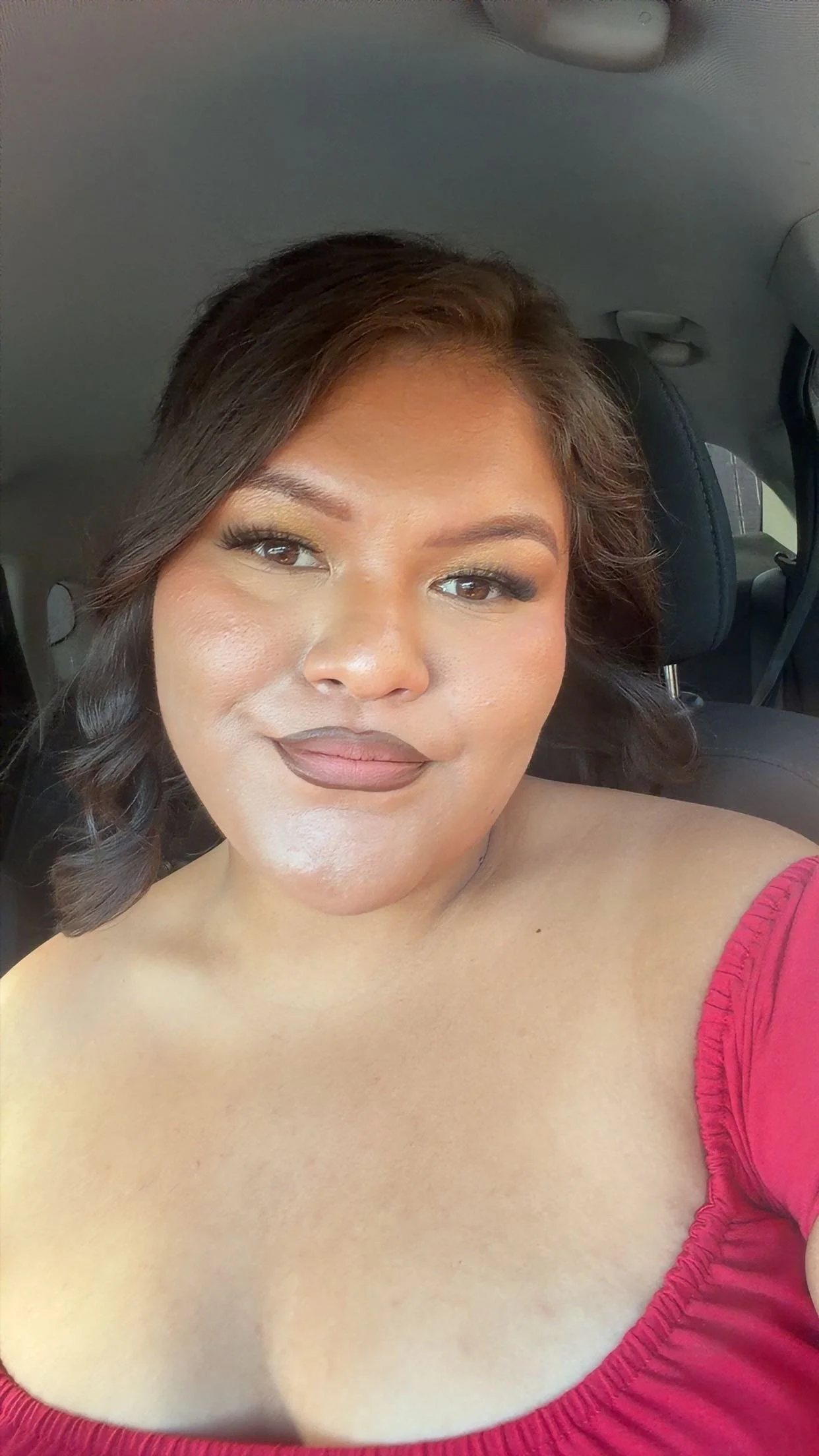 Close-up selfie of a smiling woman with shoulder-length brown hair and makeup, seated in a car wearing a pink off-the-shoulder top.