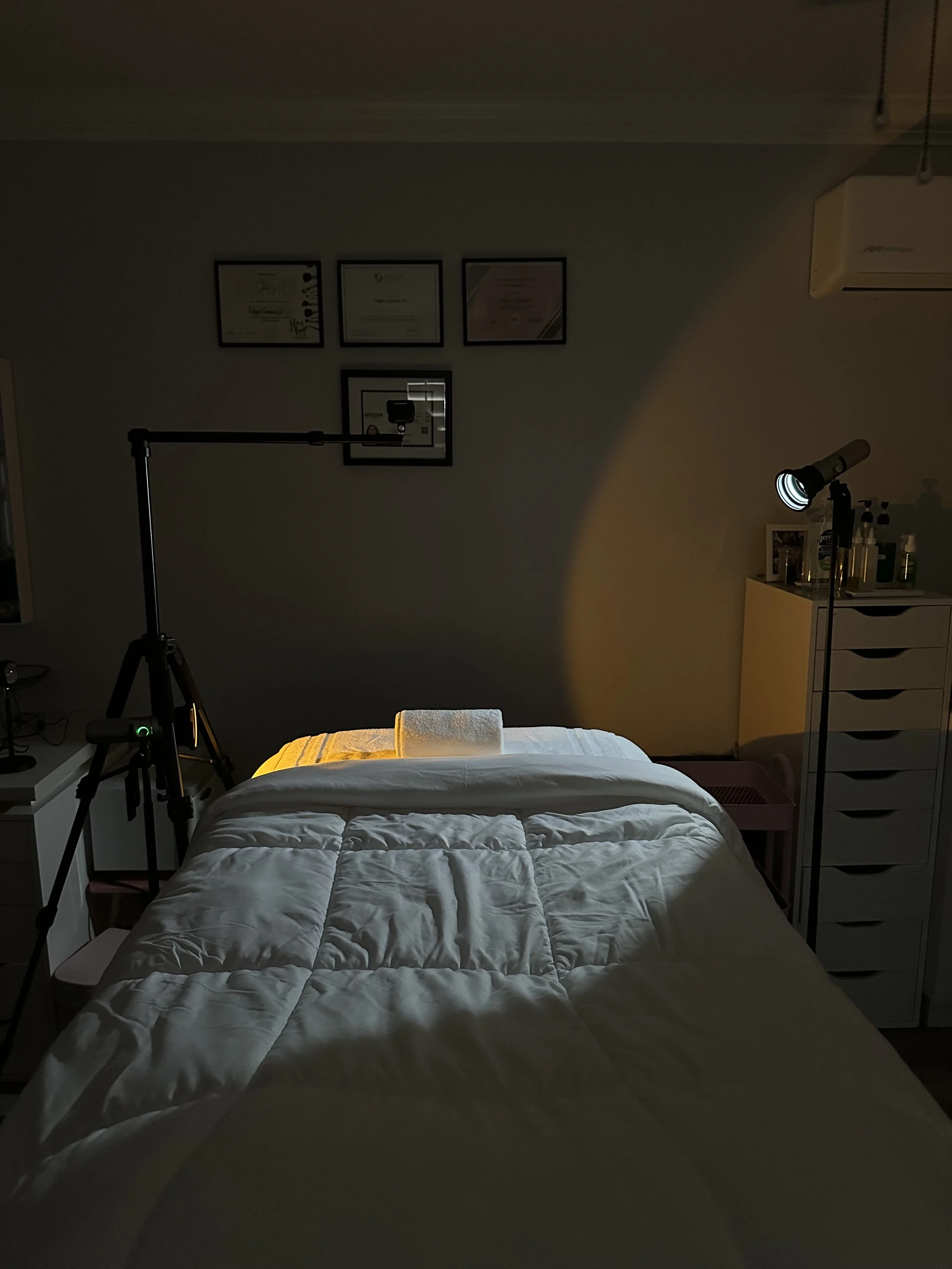 Dimly lit bedroom with a neatly made bed, framed certificates, a tripod-mounted camera, spotlight, air conditioner, and storage drawers.