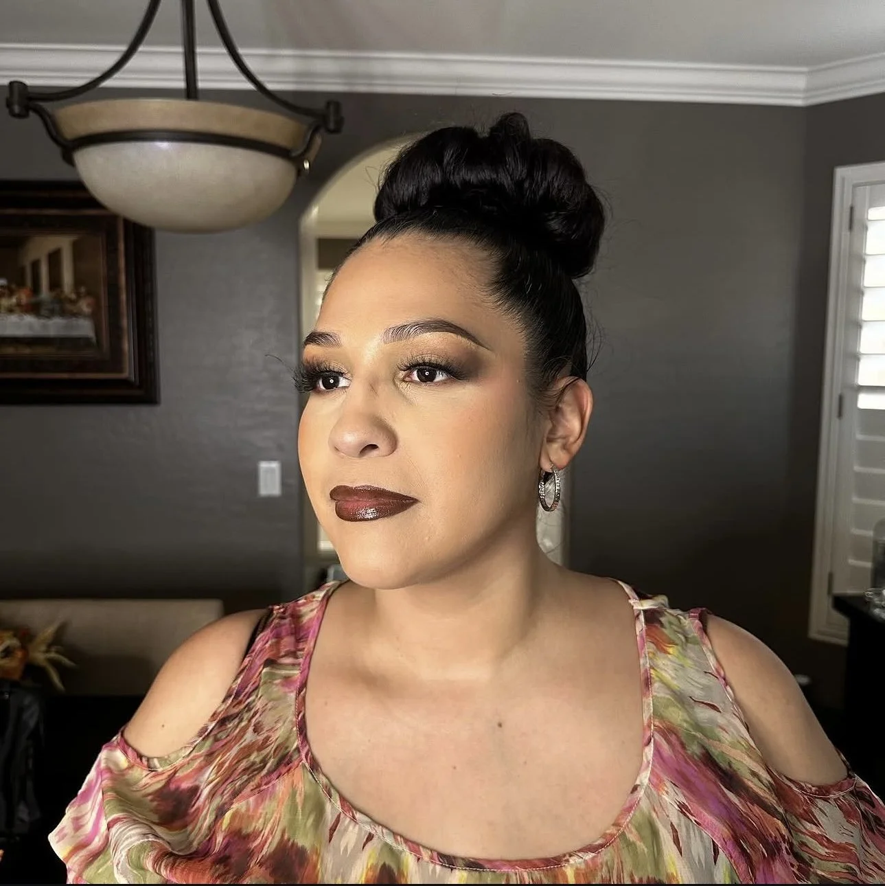 Woman with a high bun, bold makeup, hoop earrings, and a colorful cold-shoulder dress posing indoors