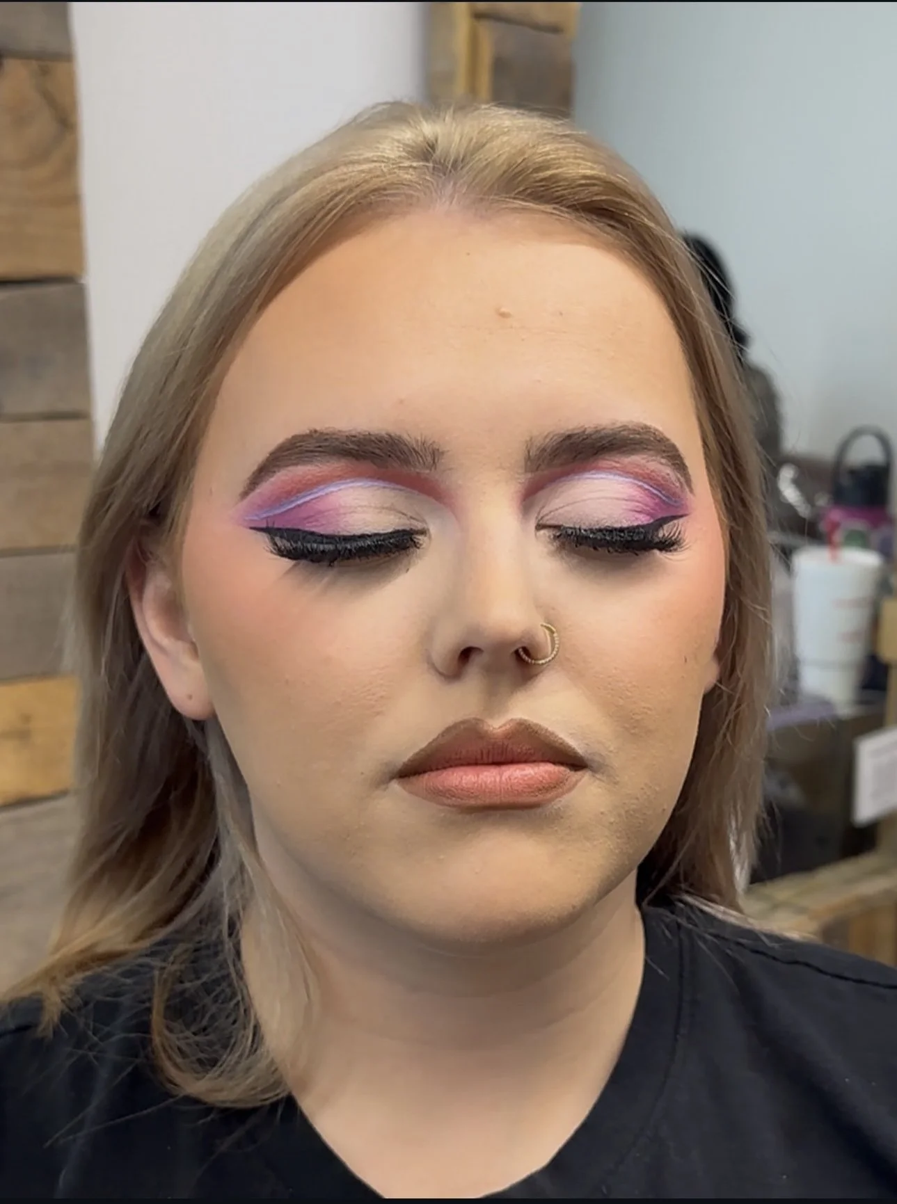 Close-up portrait of a woman with bold pink and purple winged eye makeup, false eyelashes, contoured makeup, and a gold nose ring