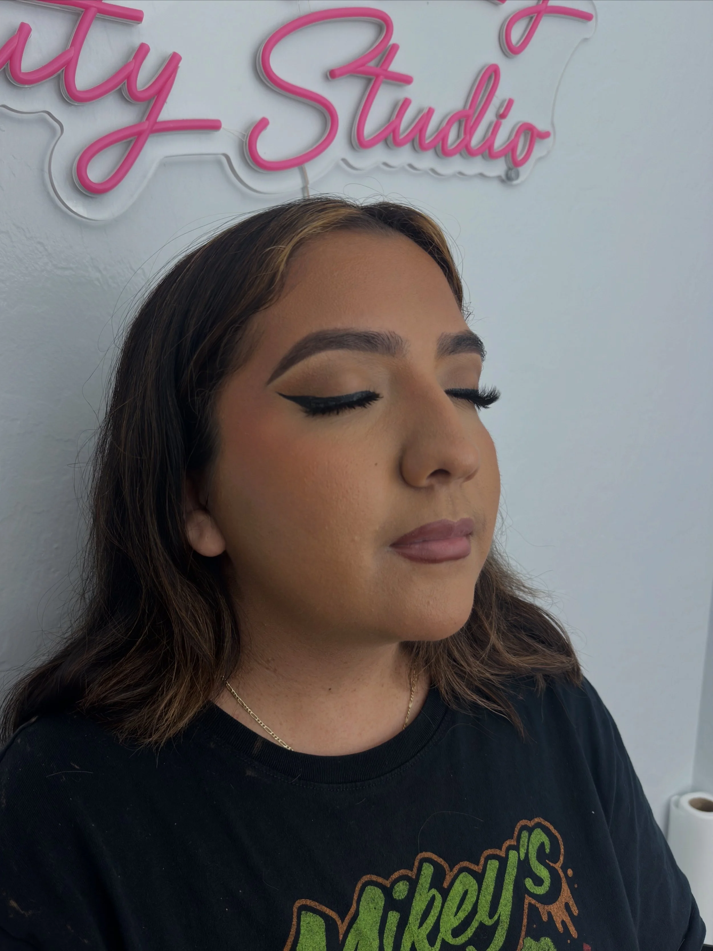 Close-up portrait of a woman with winged eyeliner and eyelash extensions, photographed beneath a pink "Edgar Skin Studio" sign.