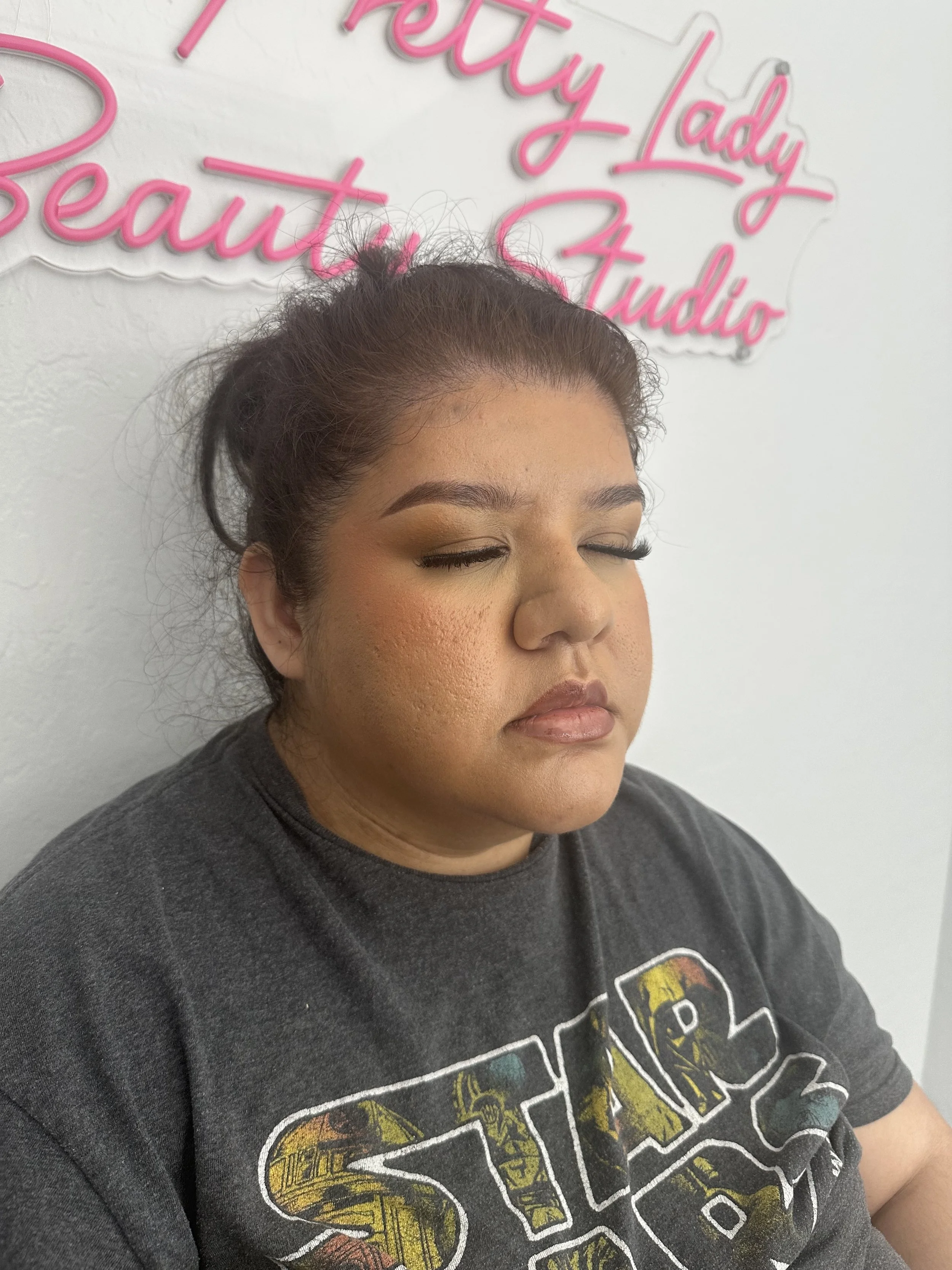 Close-up portrait of a woman with closed eyes and long eyelashes, wearing a black graphic T-shirt in front of pink cursive wall lettering.