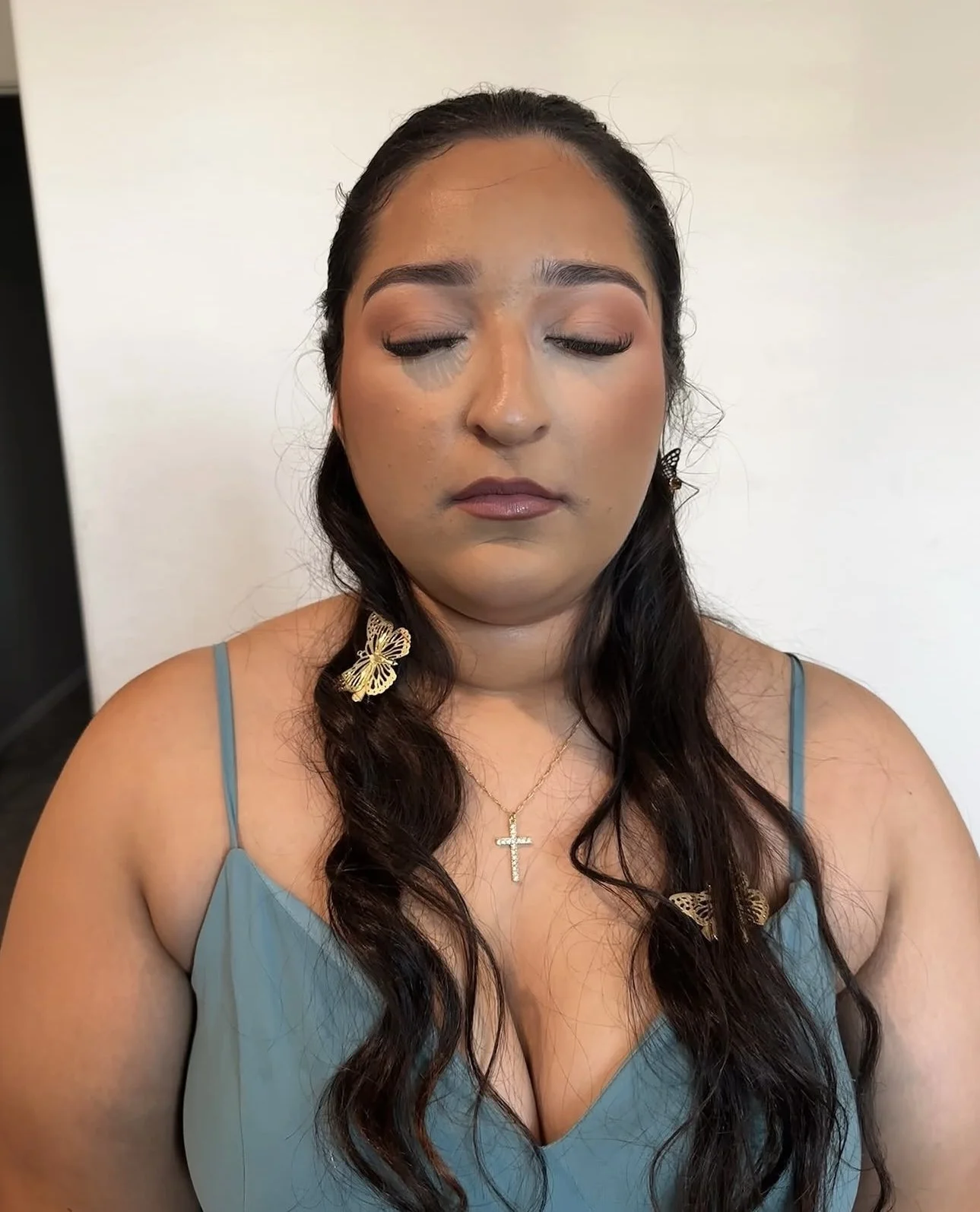 Portrait of a woman with closed eyes, long dark wavy hair, butterfly hair clips, a cross necklace, and a blue camisole