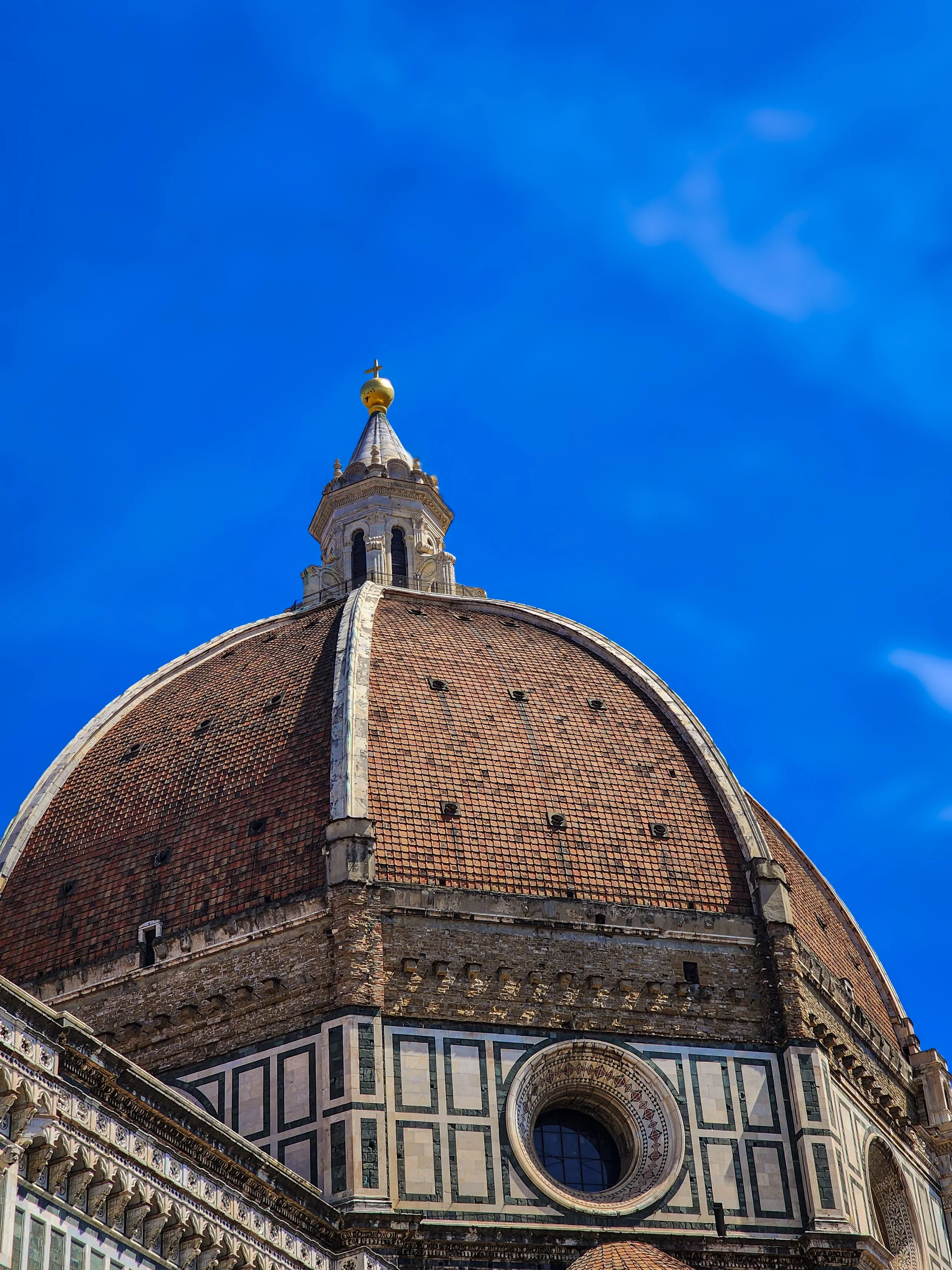 The dome of the Florence Cathedral (Cattedrale di Santa Maria del Fiore) in Italy, with a golden cross at the top, set against a bright blue sky with a few wispy clouds.