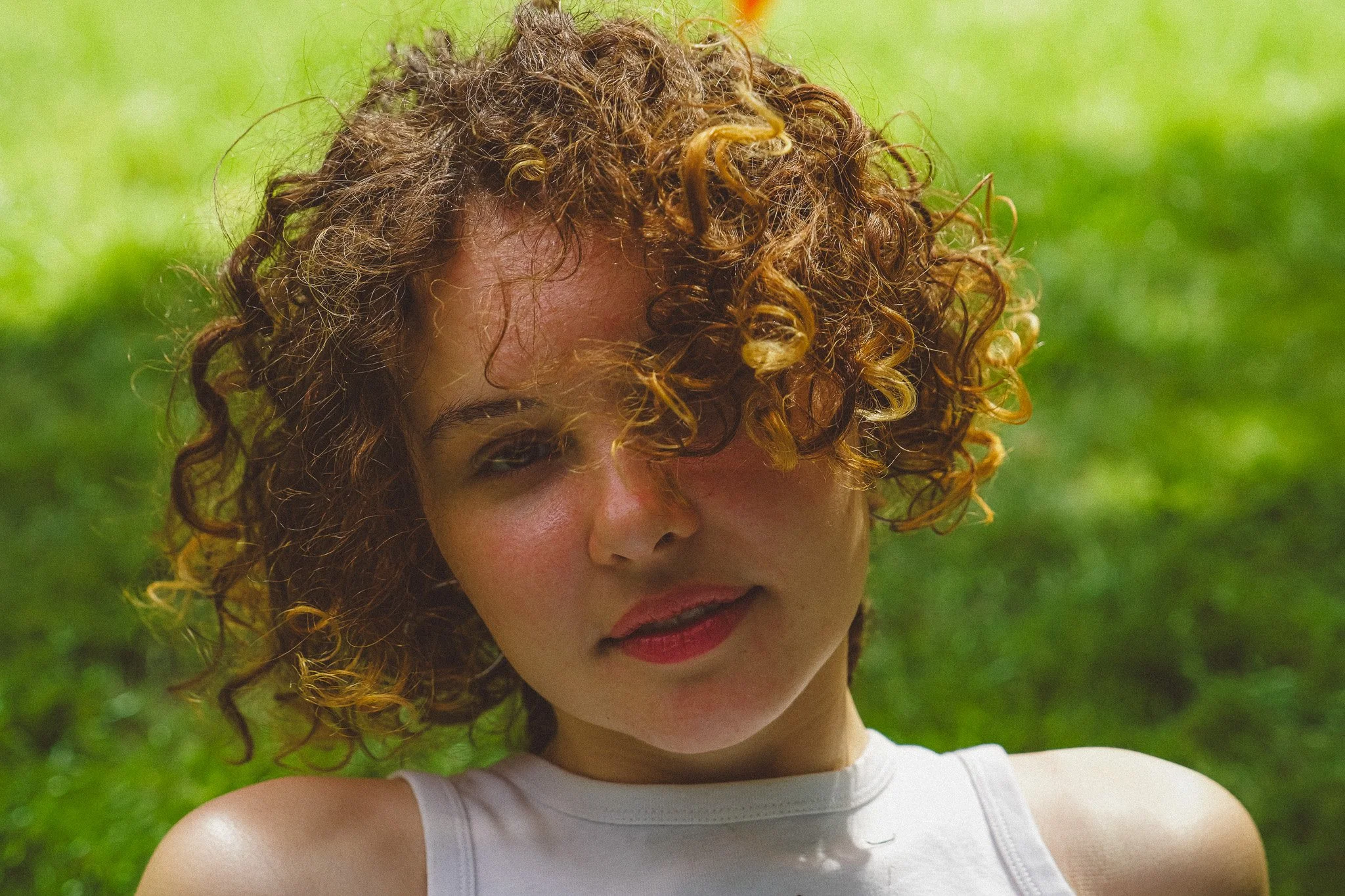 Close-up of a woman with curly red hair, green eyes, and wearing a white sleeveless top standing outside with a blurred green background.