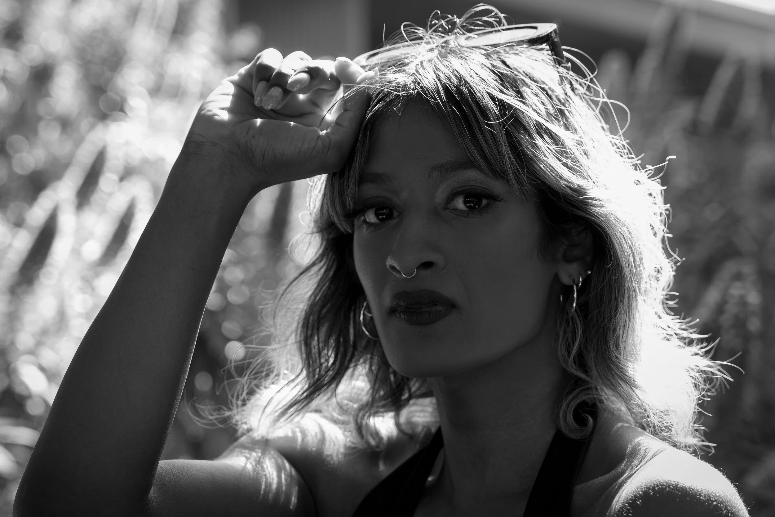 A black and white photo of a woman with light-colored hair, wearing earrings and a top, looking directly at the camera, with her right hand holding glasses on top of her head in an outdoor setting with a blurred background.