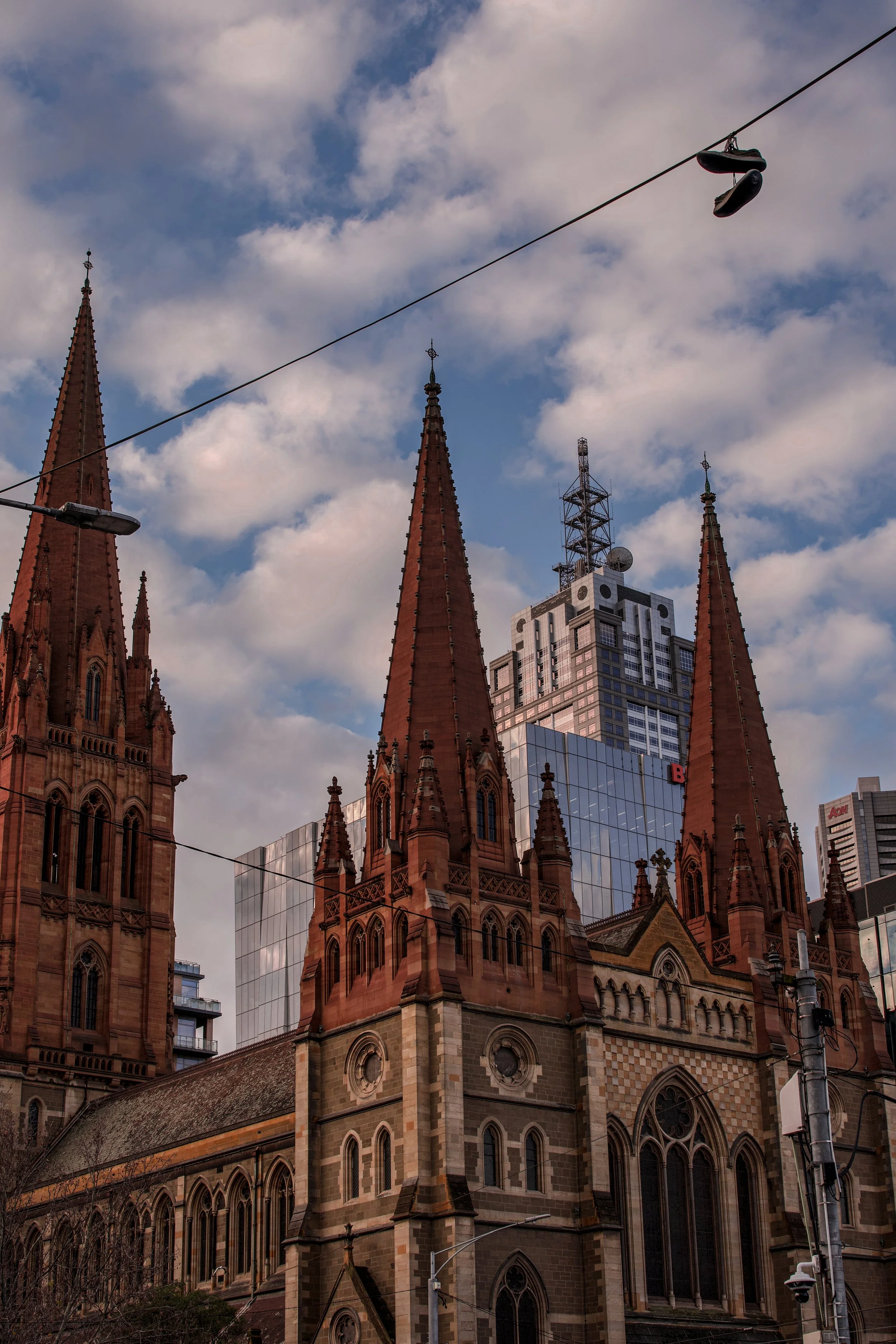 A historic church with tall, pointed spires made of red brick, set against a backdrop of modern glass high-rise buildings and a partly cloudy sky. A pair of shoes hang from a wire above the street.