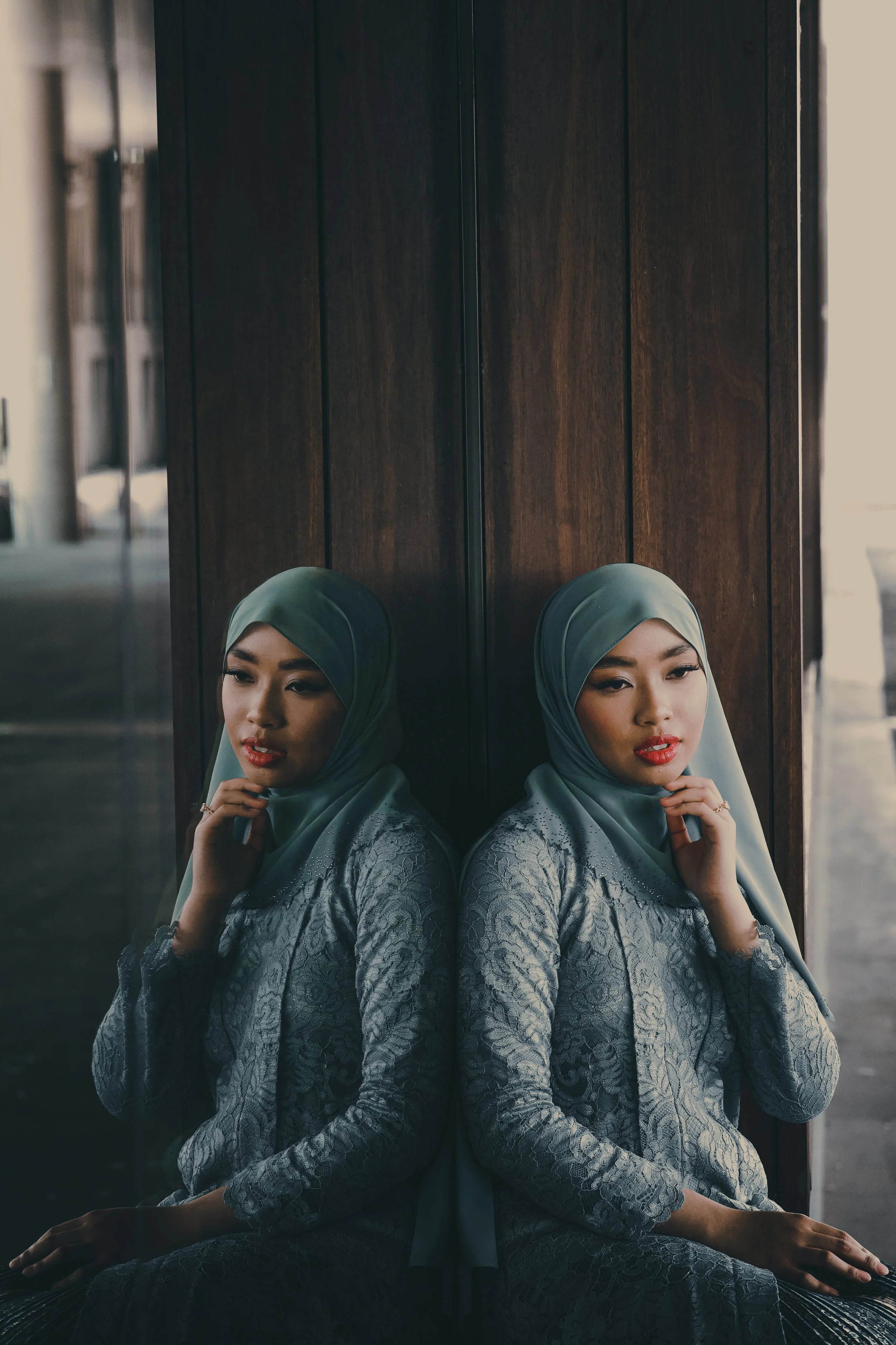 A woman in a silver dress and gray hijab sitting with her back against a wall, reflected in a mirror. She has a thoughtful expression, with her hand near her chin.