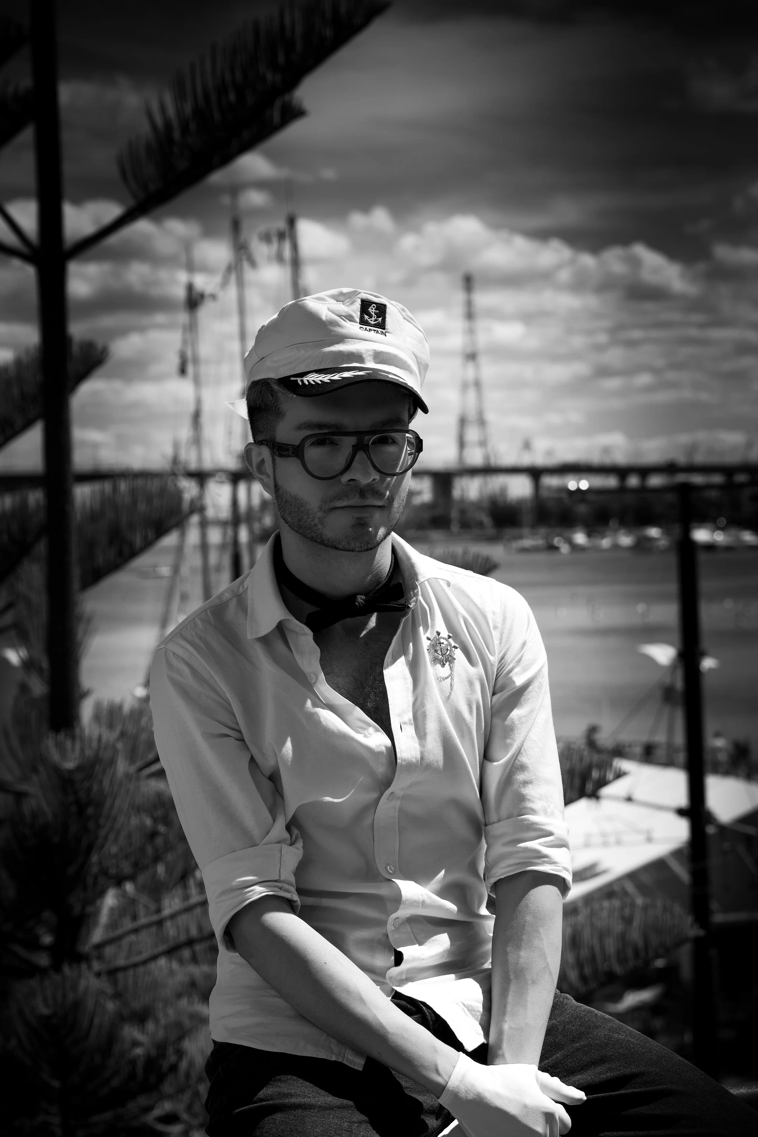 Black and white photo of a man in sunglasses and a captain hat, sitting outdoors with a bridge and power lines in the background.