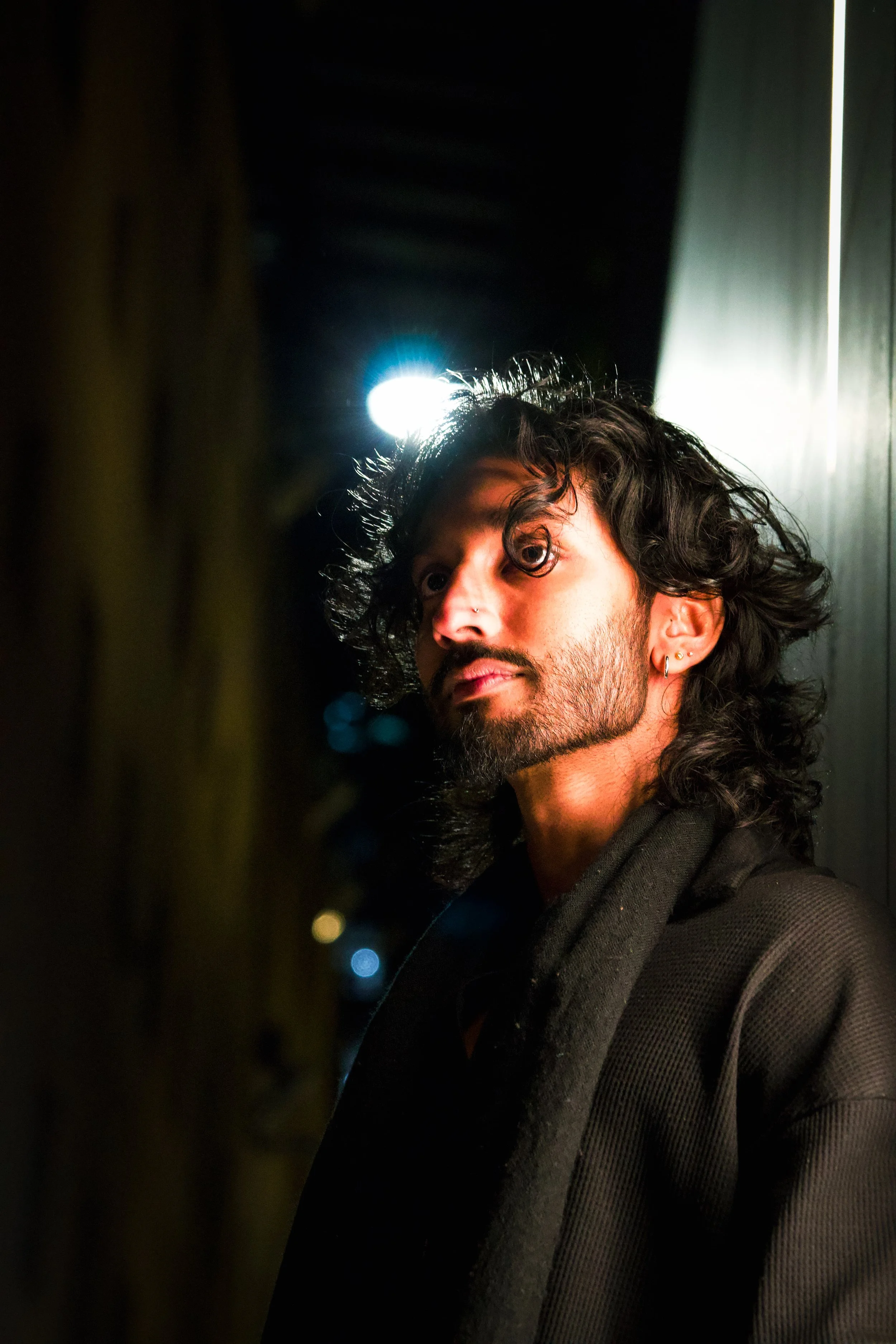 A man with long curly black hair, wearing a black hoodie, looking into a camera in a dimly lit space with bright lights behind him.