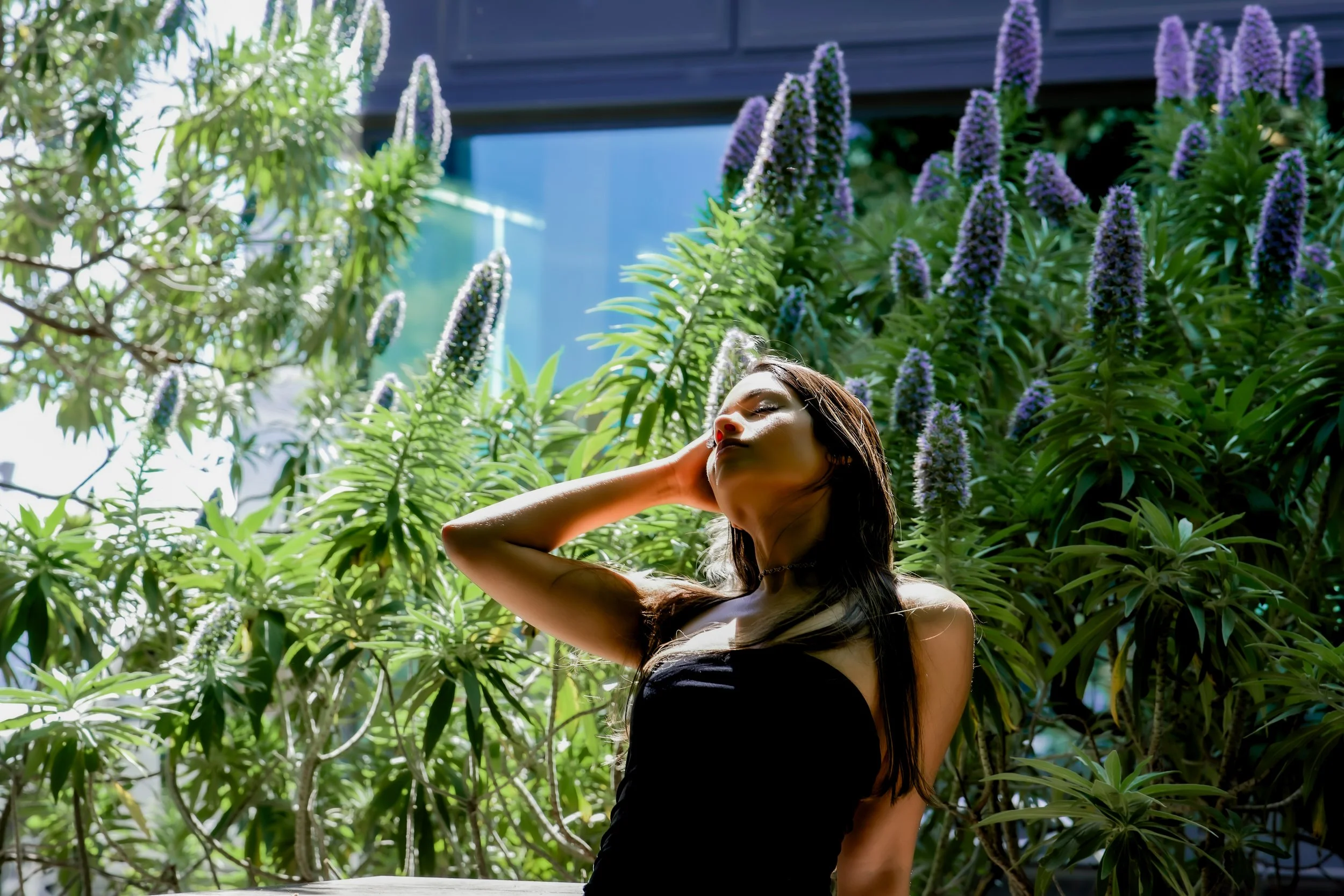 A woman with long dark hair wearing a black dress, standing outdoors among lush green plants and purple flowers, with her eyes closed and one hand touching her face.