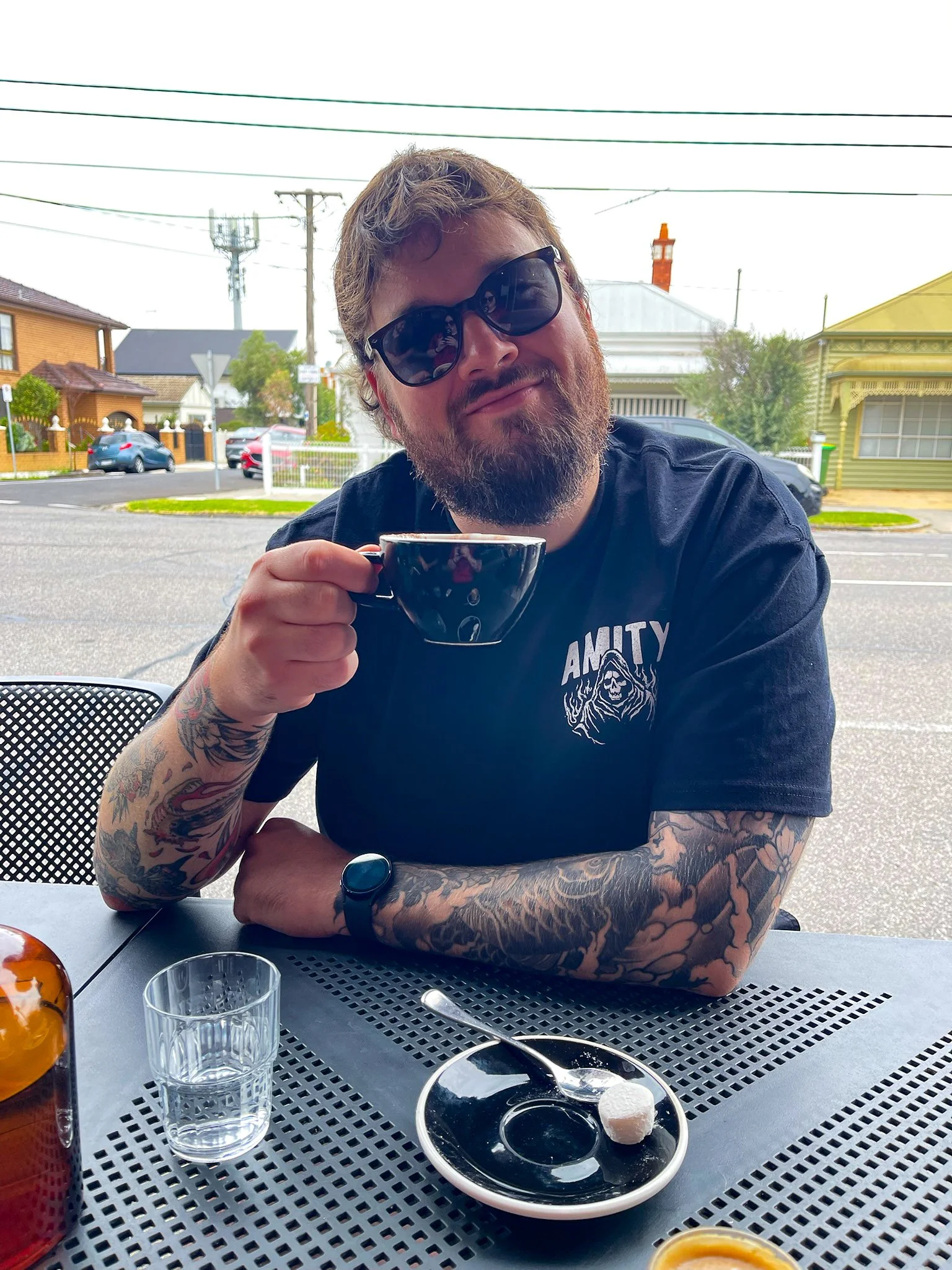 A man with tattoos on his arms, wearing sunglasses and a black T-shirt with a graphic design, sitting at an outdoor table holding a black coffee cup. The table has a glass of water, a spoon, a small plate with a white dessert ball, and a container of orange sauce. The background shows a residential neighborhood with houses, trees, and power lines.