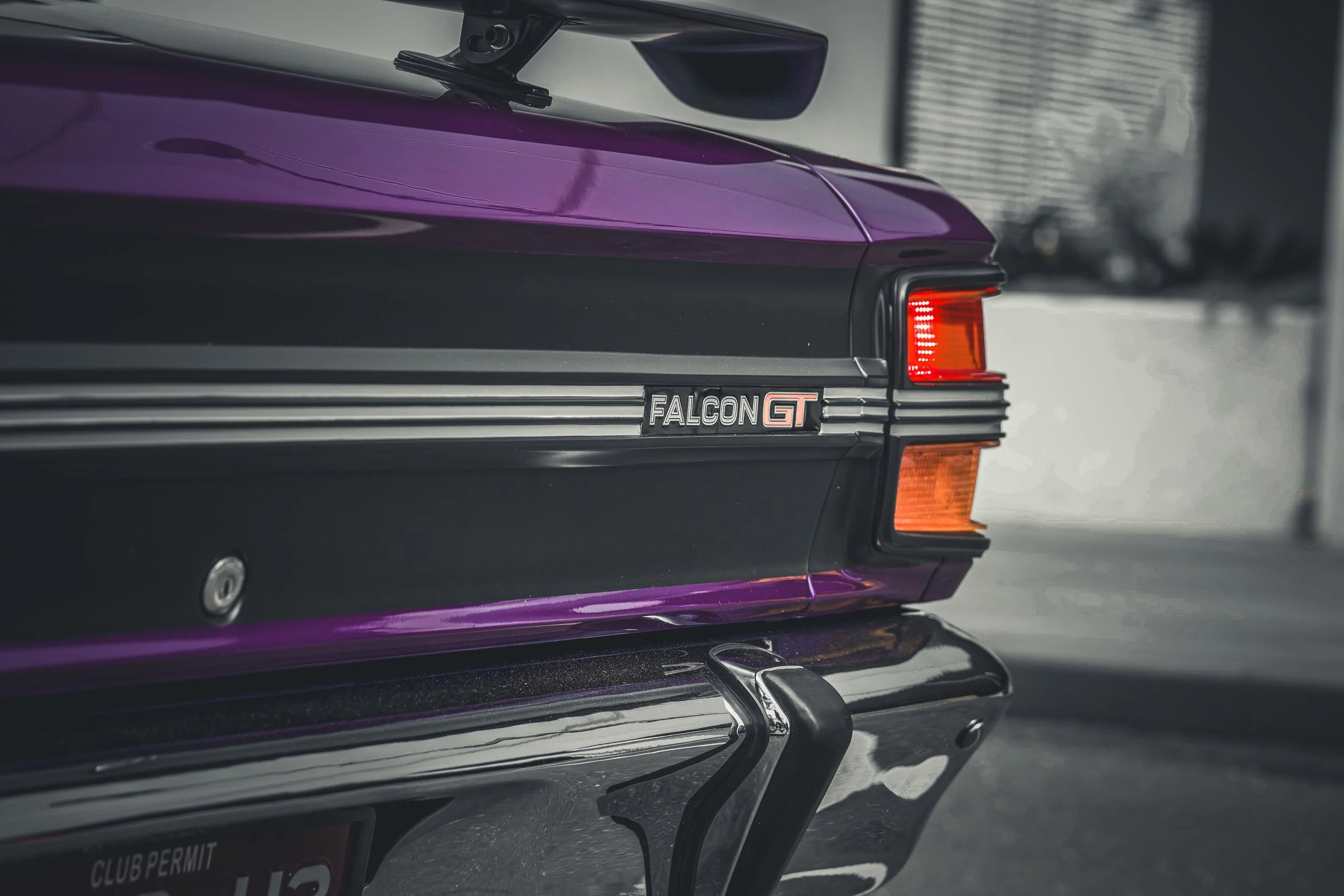 Close-up of the rear of a vintage Pontiac Trans Am Pontiac Firebird, showing the purple paint, tail light, and 'Falcon GT' emblem, with a cityscape in the background.