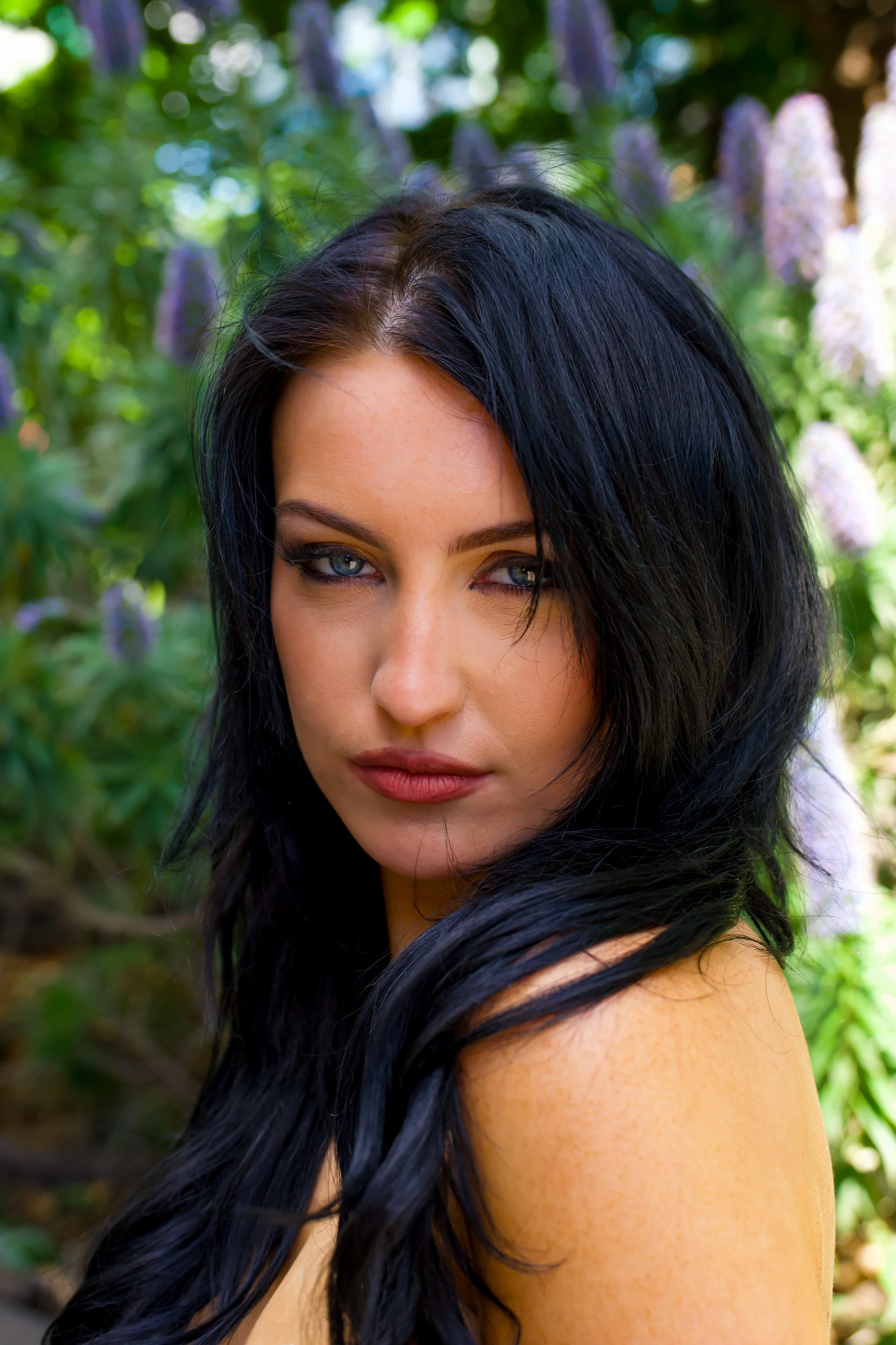 Close-up of a woman with long black hair and blue eyes, outdoors with blurred green foliage and purple flowers in the background.