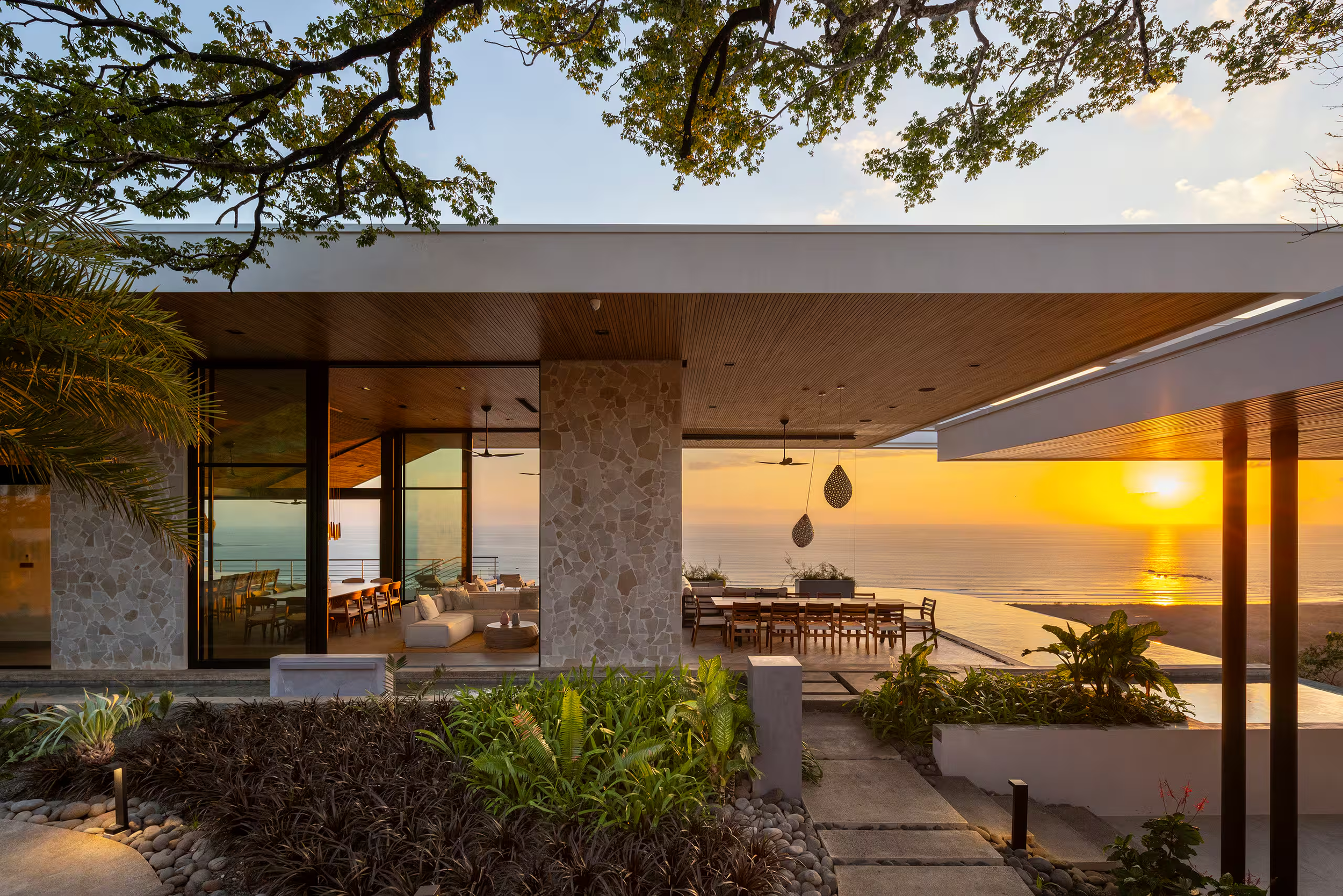 Modern house overlooking the ocean at sunset with outdoor dining and living areas, surrounded by lush greenery and decorative stones.