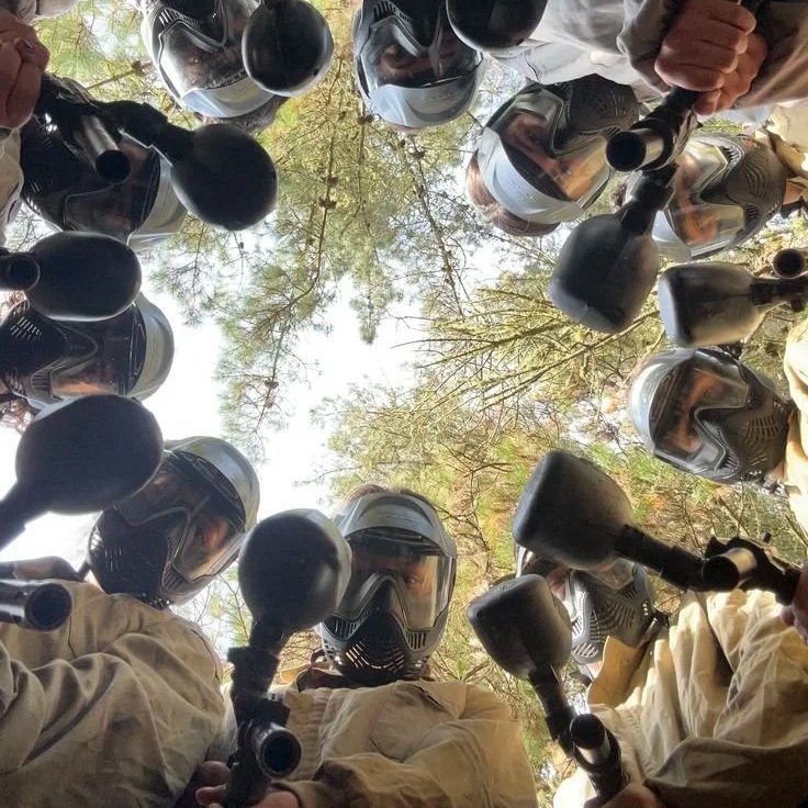 Paintball players in protective gear and helmets forming a circle outdoors, looking down at the camera.